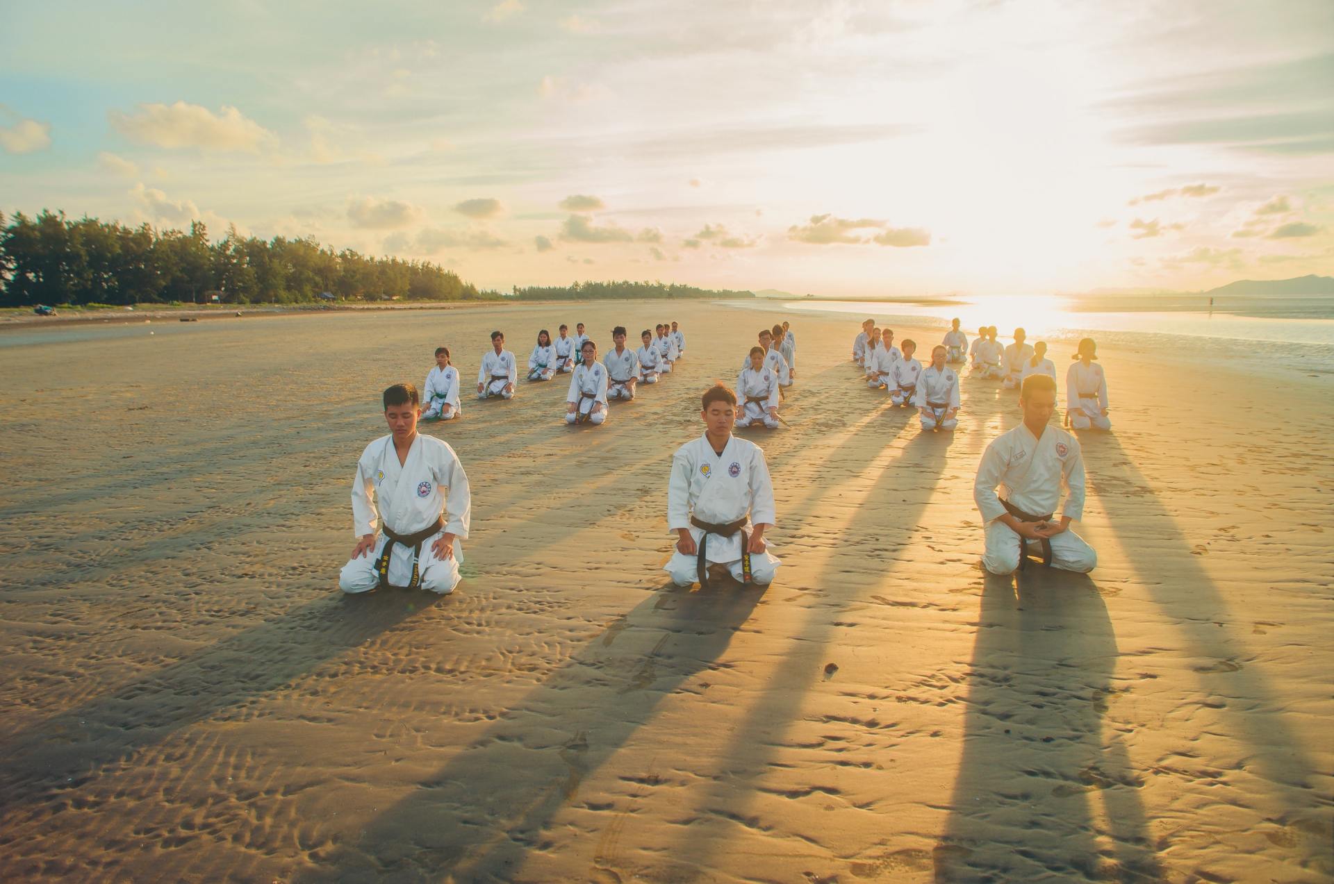 Karate practitioners in white gis kneel on a beach at sunrise.