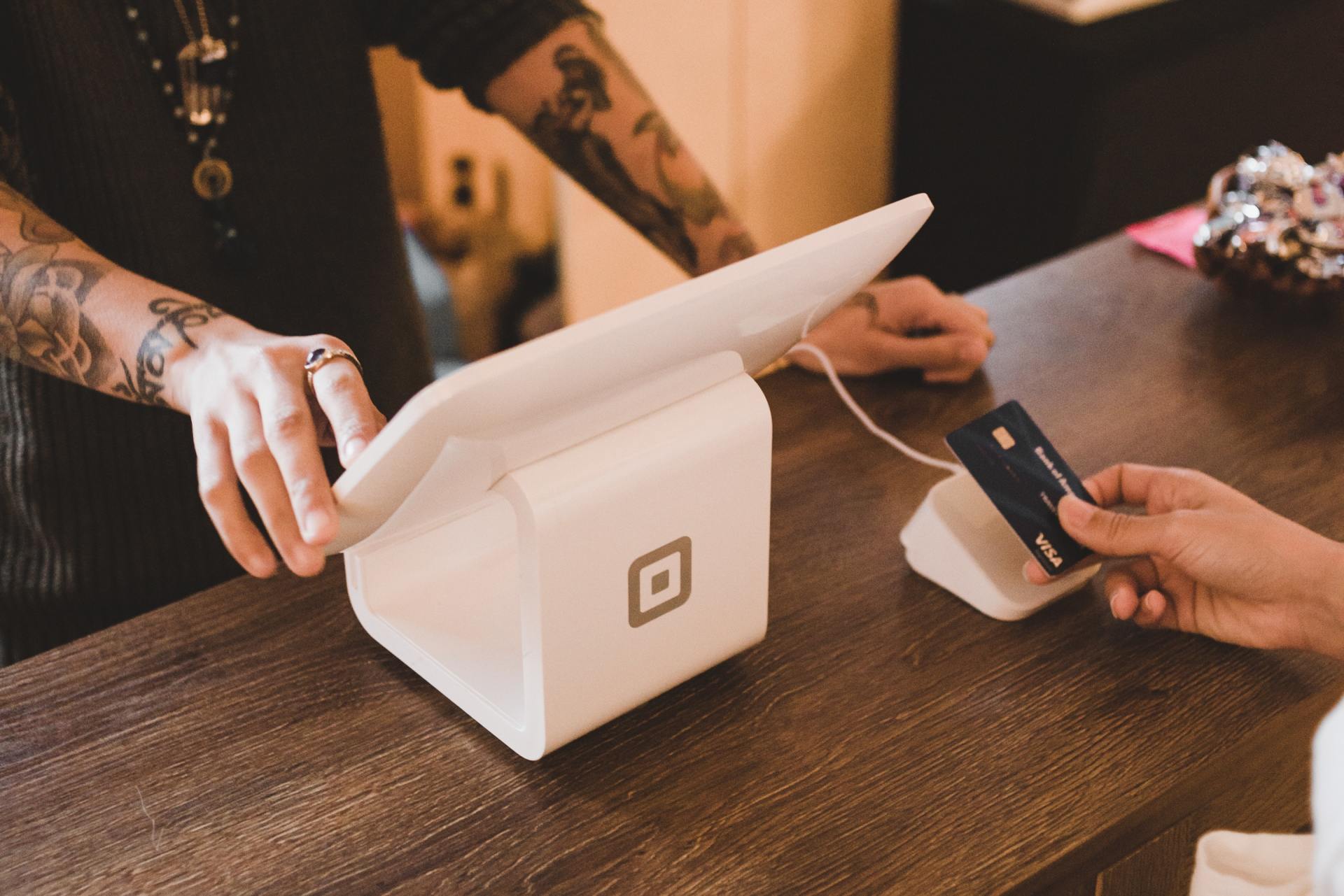 Hands using a card terminal and payment card over a white checkout box on a wooden counter