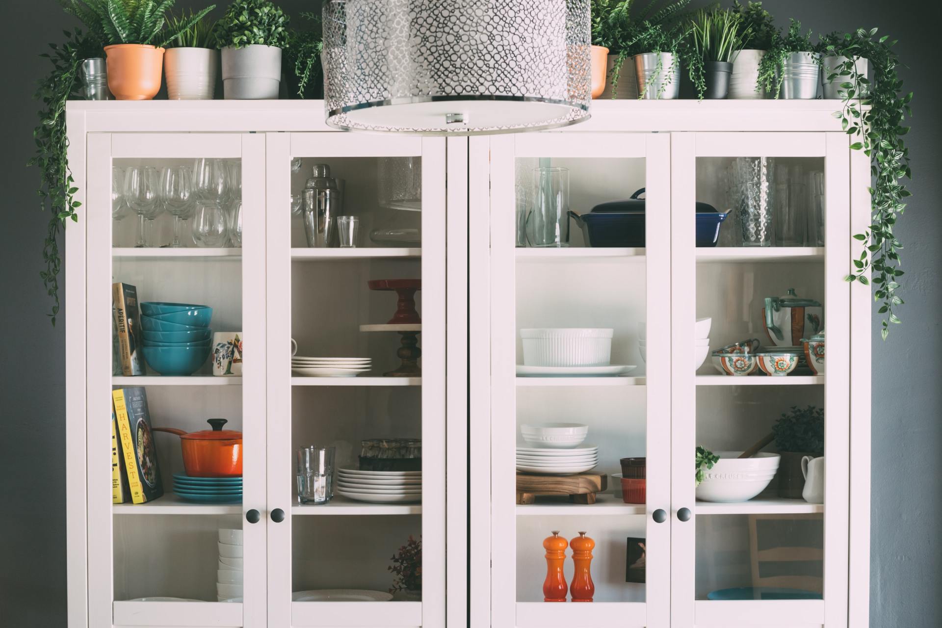 White cabinet with glass doors, filled with dishes and decor, plants on top, and hanging vines.