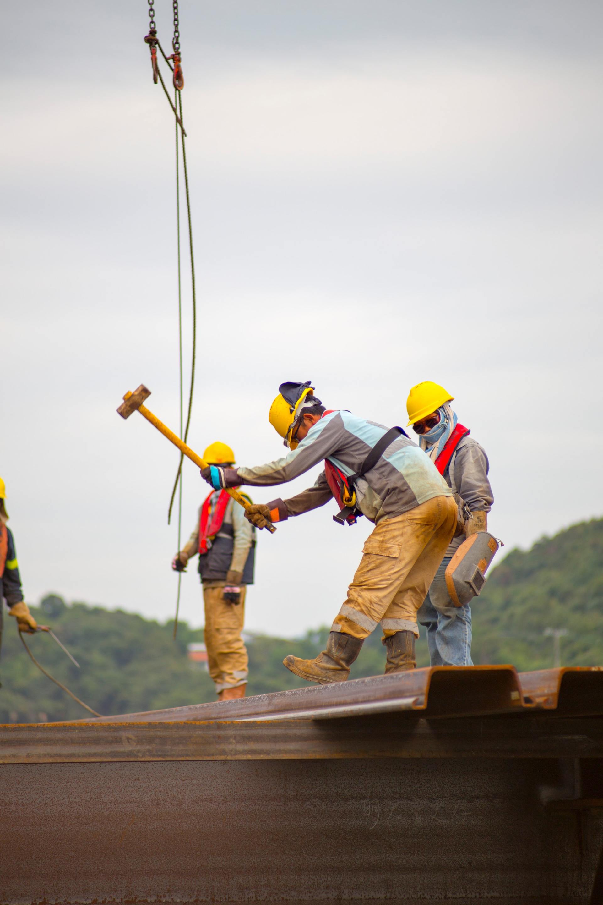 Construction workers hammering on steel beam, wearing helmets and safety gear, cloudy sky background.