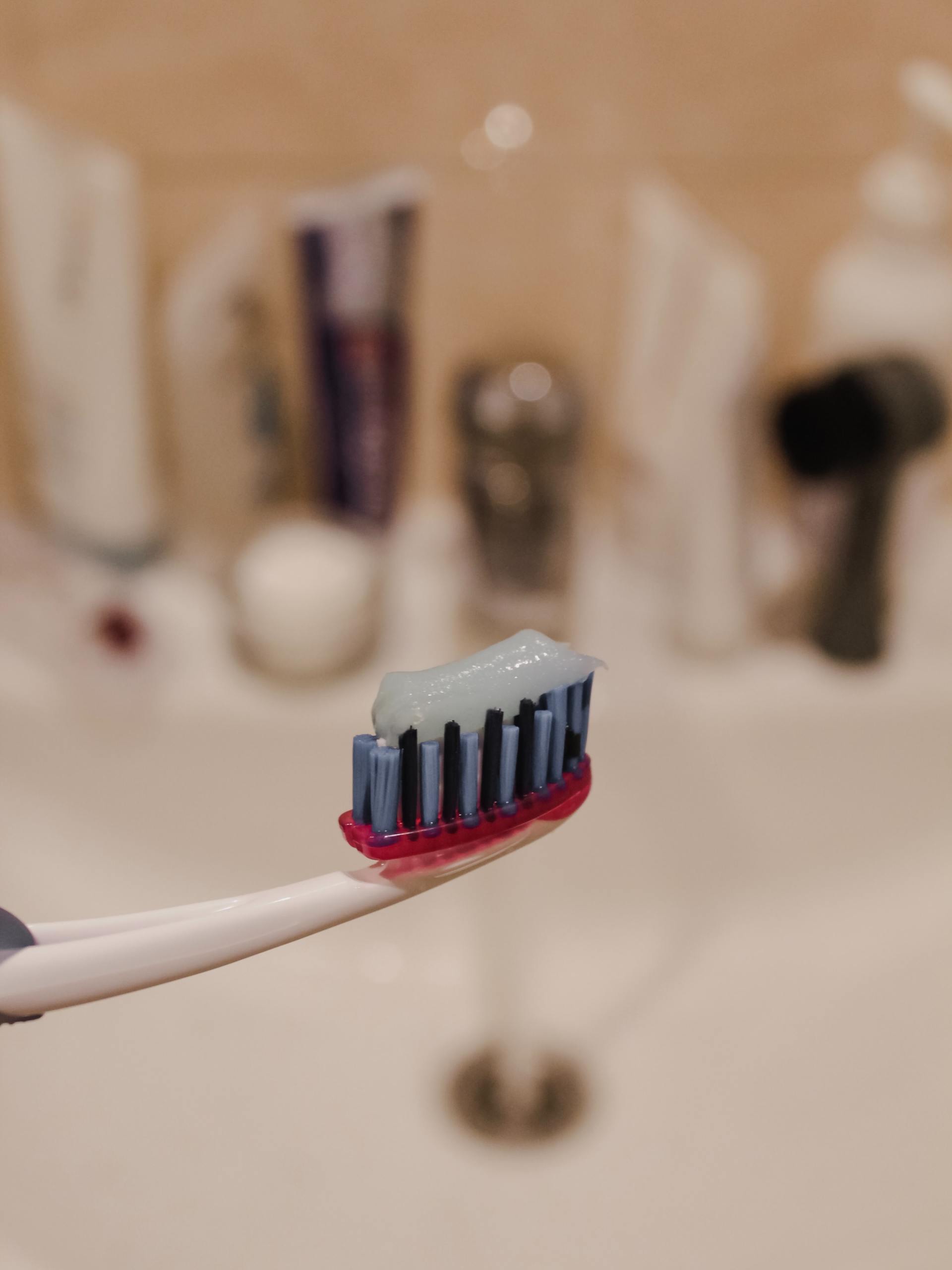 Toothbrush with toothpaste, held in front of a bathroom sink, other toiletries blurred in the background.