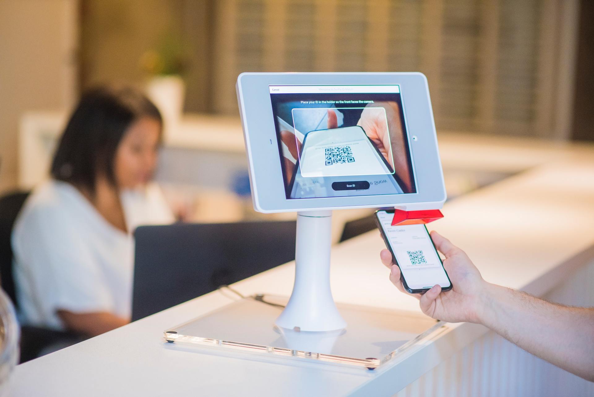 A person scans a QR code on their phone with a kiosk, another person works on a laptop in the background.