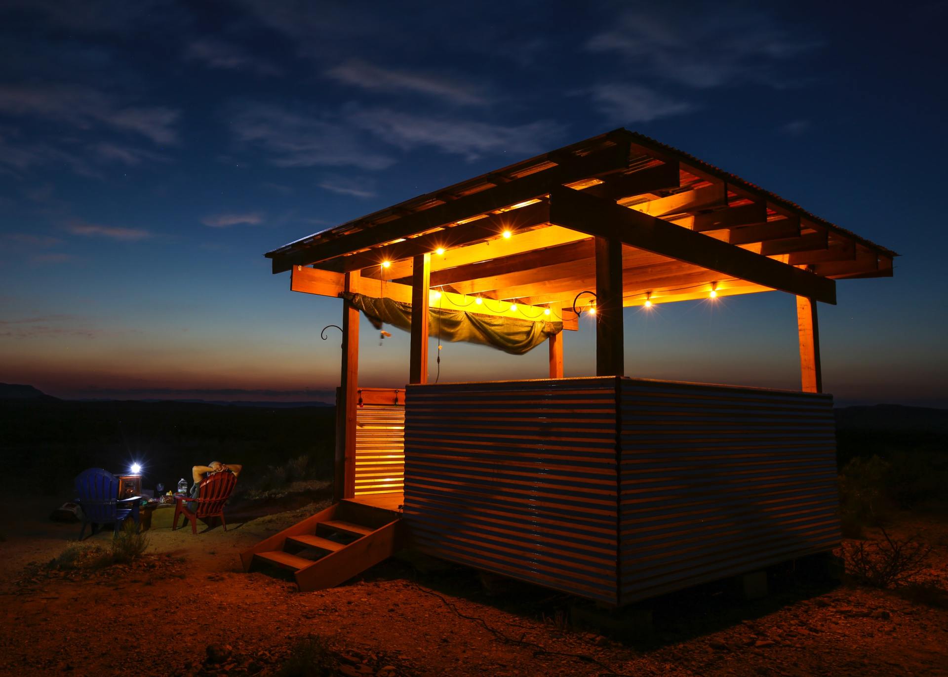 Outdoor wooden structure with string lights, hammock inside, and camping chairs at dusk.