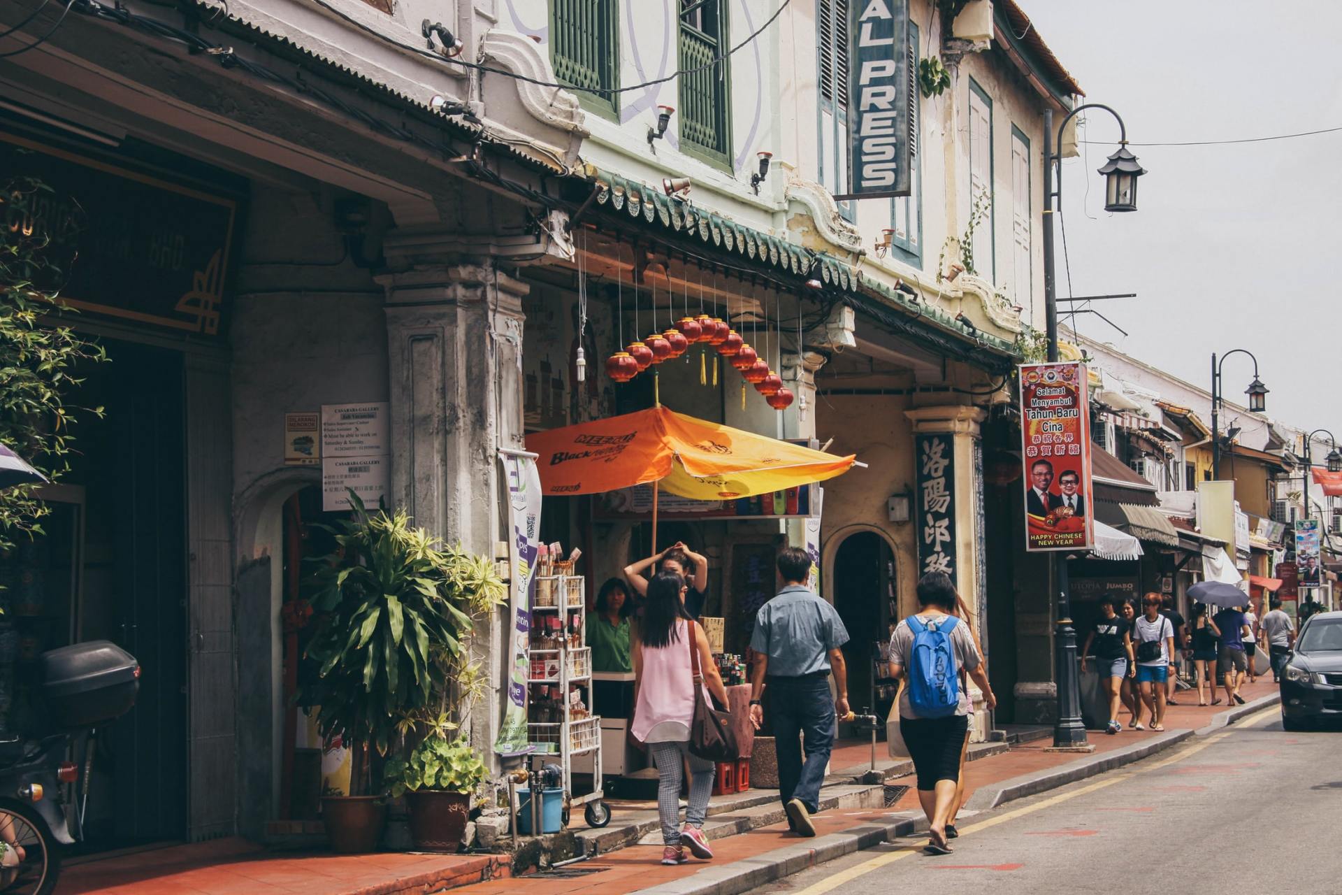 People walking past colorful shops and cafés on a narrow street with awnings and potted plants.