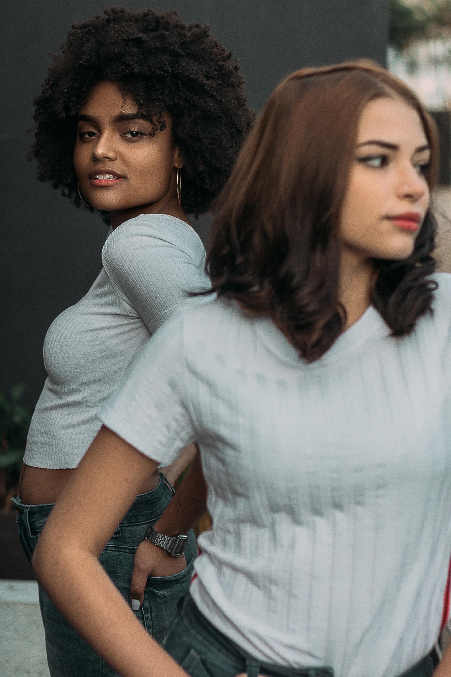 Two young people posing outdoors in white tops, jeans, one with curly hair, the other with straight.