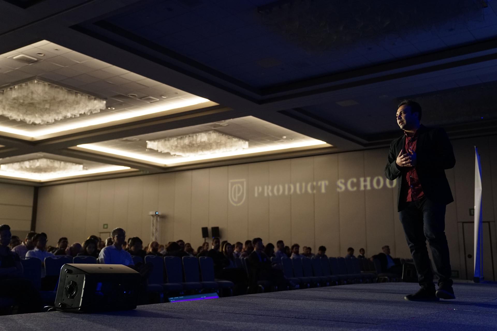 Como transformar um curso presencial em on-line? Um homem está no palco em frente a uma multidão em uma conferência de uma escola de produtos.