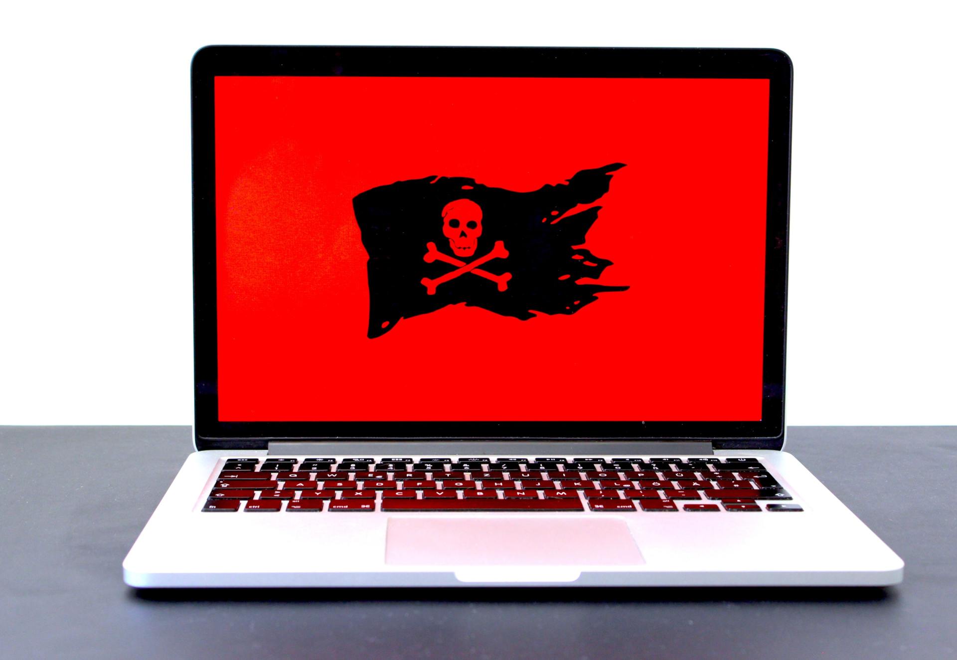 Sobre una mesa hay un ordenador portátil cuya pantalla muestra un emblema de una calavera con tibias cruzadas de color negro sobre un fondo rojo liso.