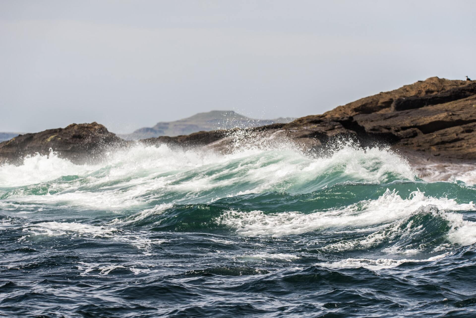 Waves crash against rocky coast; turquoise water, white foam, gray sky.
