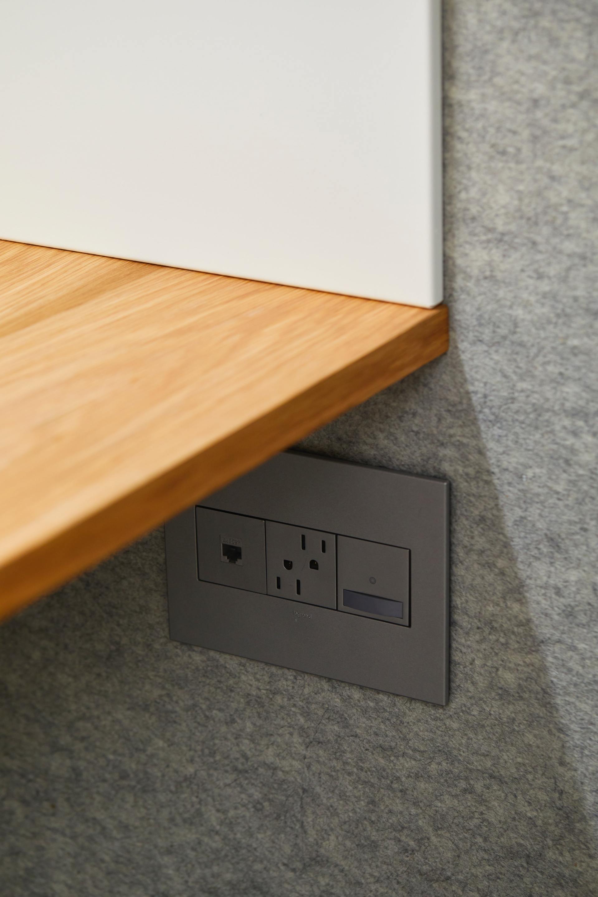 Dark gray electrical outlet panel mounted on a gray textured wall below a wooden shelf.