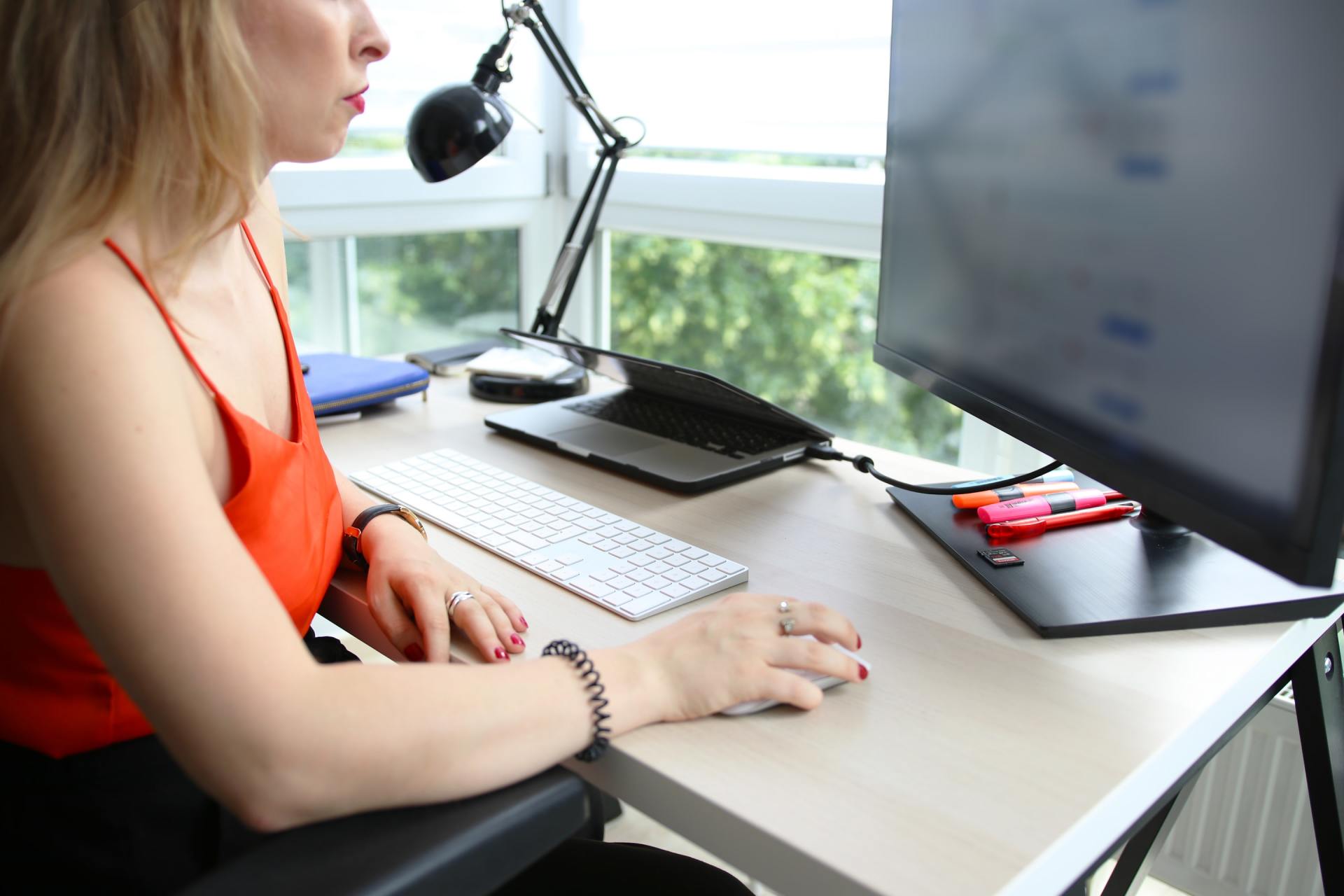 Woman typing at a desk with dual monitors, keyboard, and desk lamp by a bright window