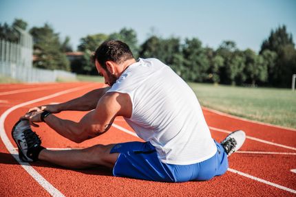 Man stretching on a red running track, reaching for his foot. Wearing white shirt, blue shorts. Sunny outdoor setting.