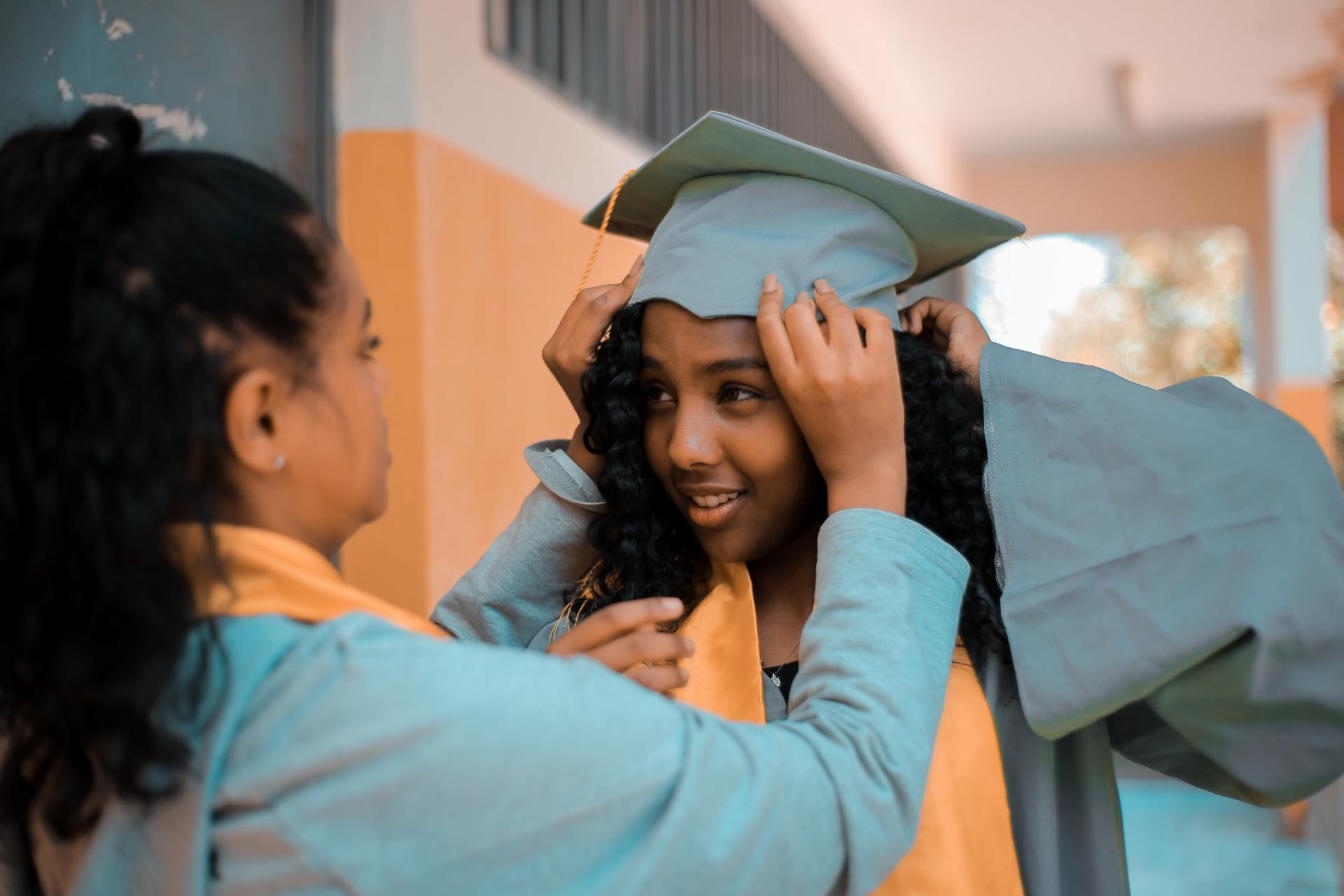 A woman is helping another woman put on her graduation cap and gown.