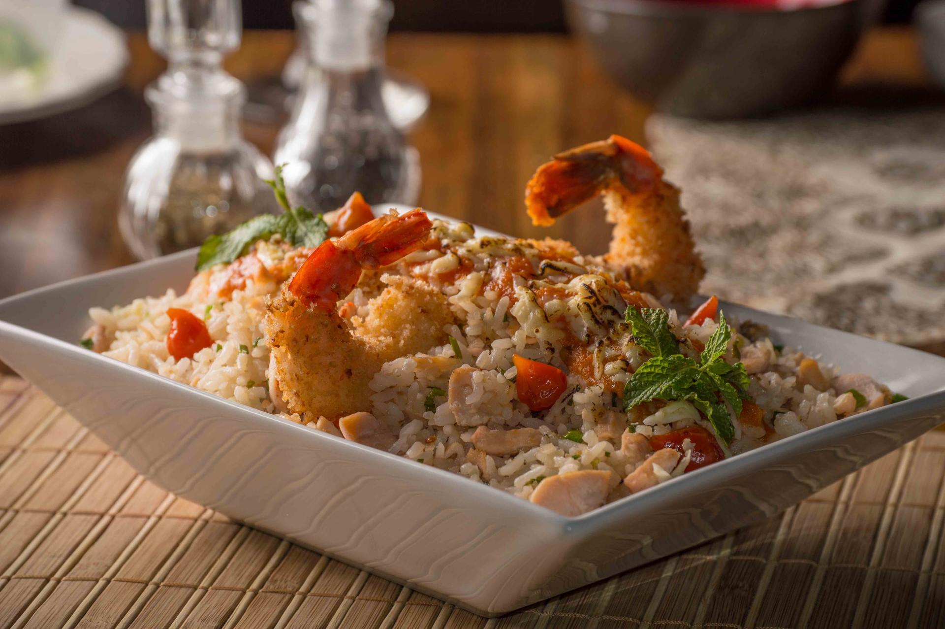 Prato de camarão com arroz, decorado com ervas, disposto sobre uma mesa, com vidros embaçados ao fundo.