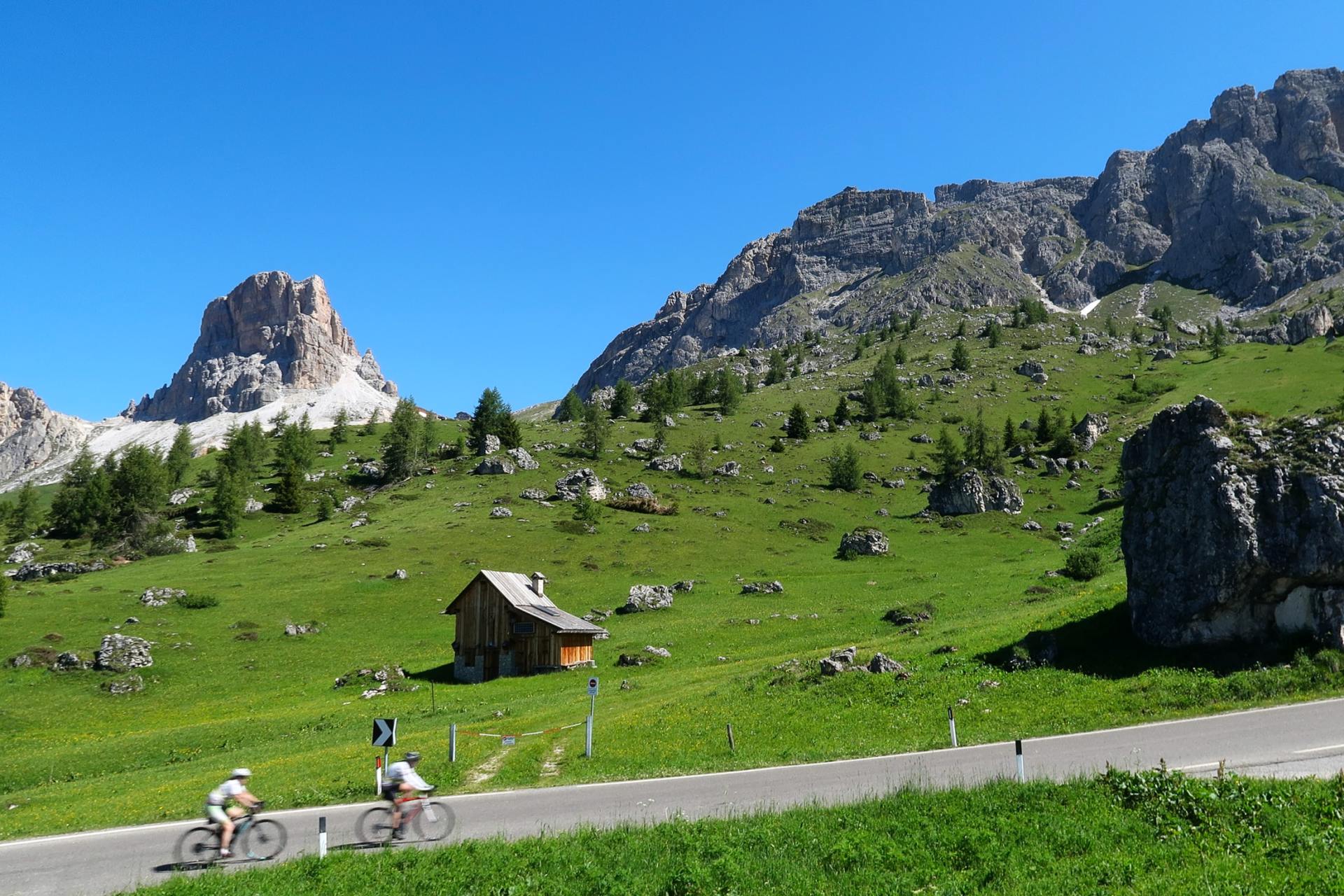 Radfahrer auf einer Bergstraße neben einer grünen Almwiese, einer Hütte und felsigen Gipfeln unter blauem Himmel