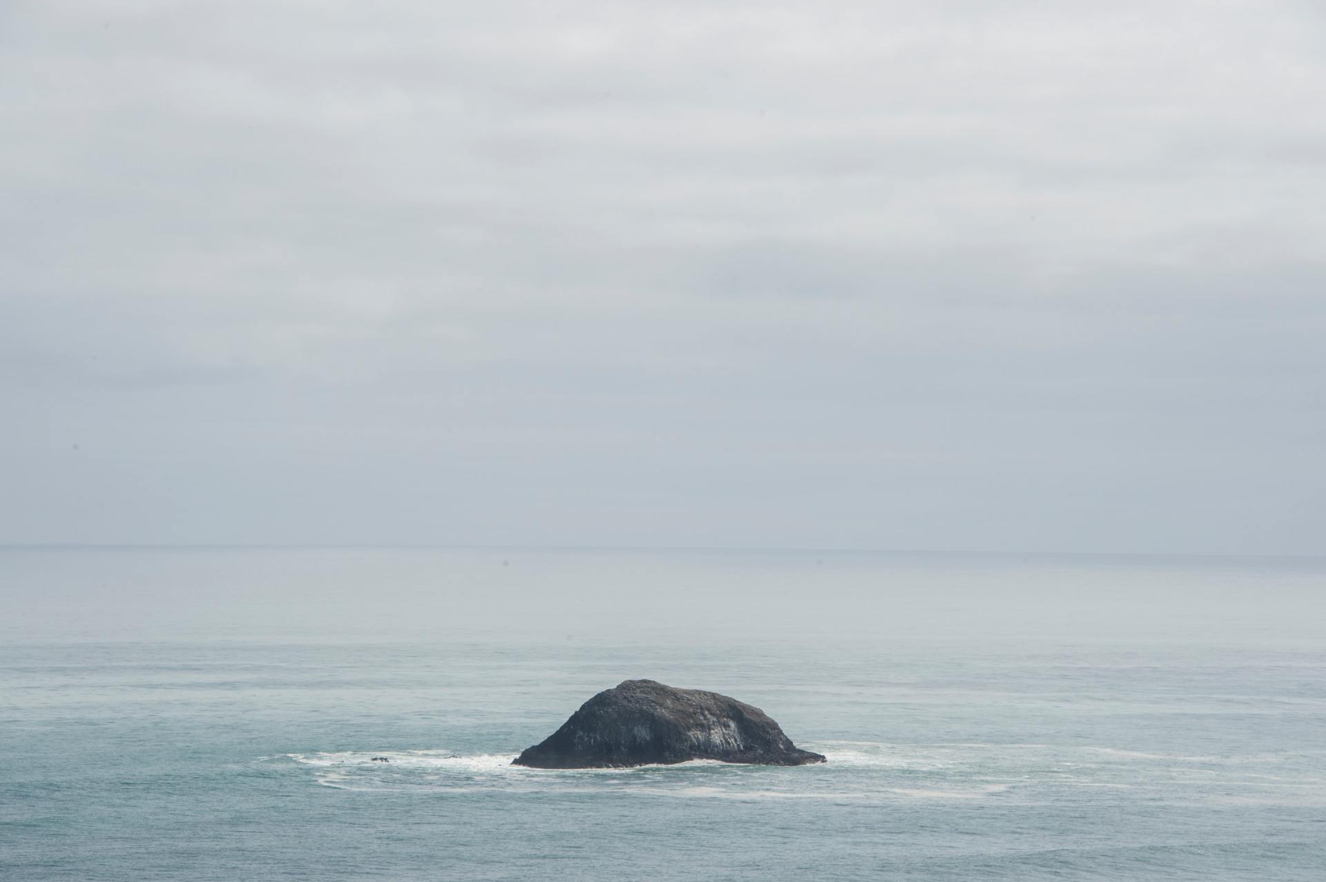 Una solitaria e scura formazione rocciosa si erge al centro di un oceano calmo e grigio, sotto un cielo nuvoloso.