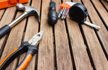 A collection of home repair tools, including a hammer, pliers, screwdriver, and tape measure, on a slatted wooden table.