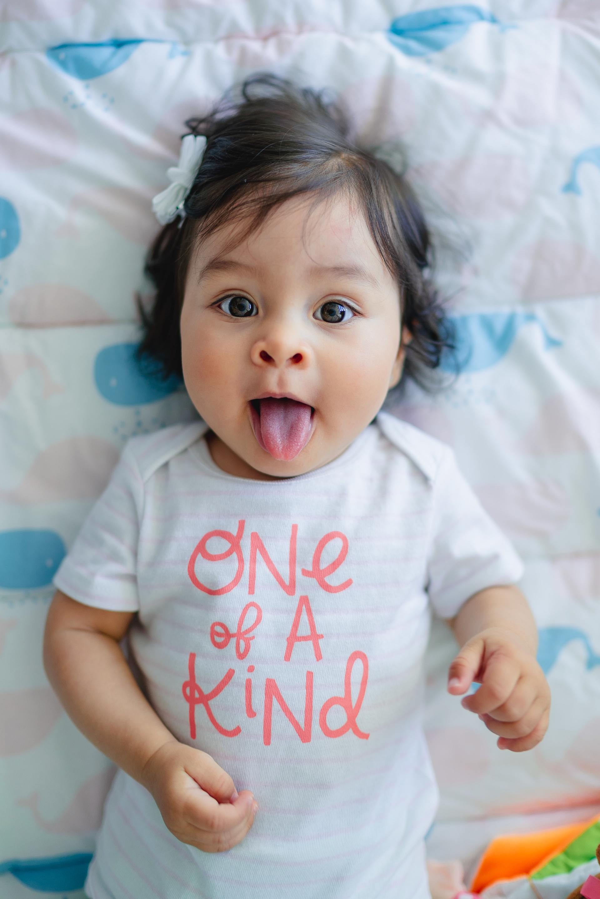 A baby girl is laying on a bed with her tongue out and wearing a one of a kind shirt.