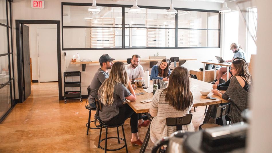 Group of people seated around a long wooden table in a brightly lit office space, engaged in a discussion.