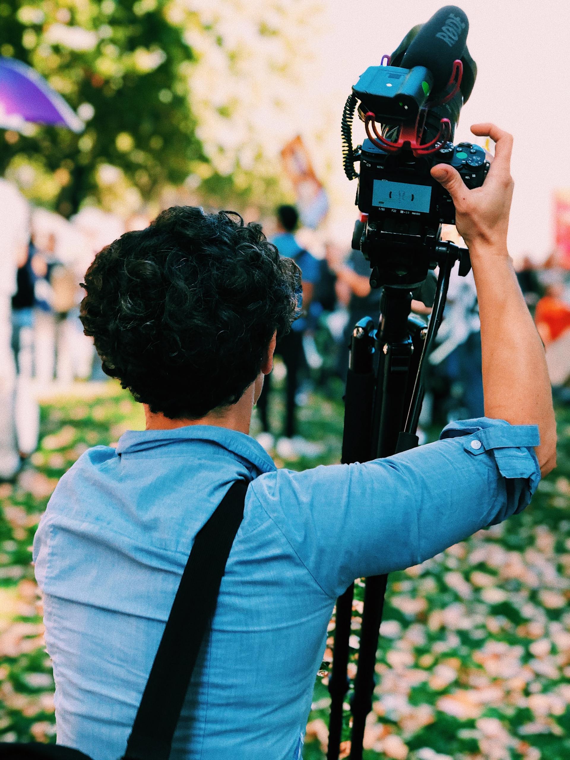 Person filming with a camera on a tripod outdoors, surrounded by people, wearing a blue shirt.