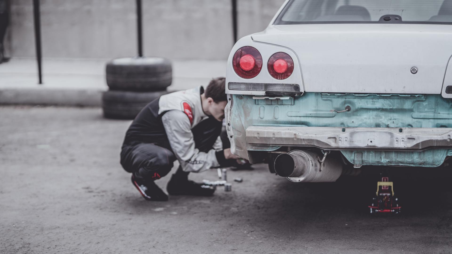 A mechanic working on the rear of a white car, crouching near the exhaust pipe; tires stacked nearby.