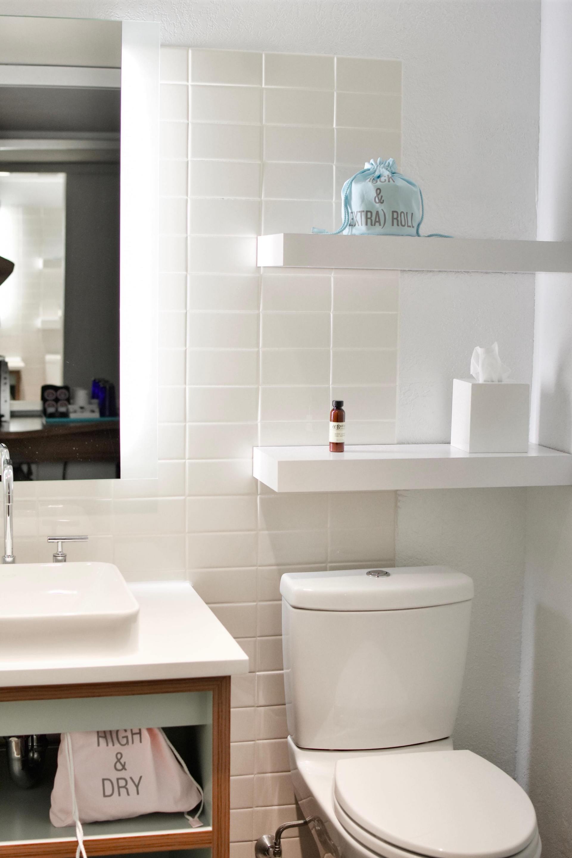 A bright bathroom with a white sink, toilet, and two floating shelves against a white tiled wall.