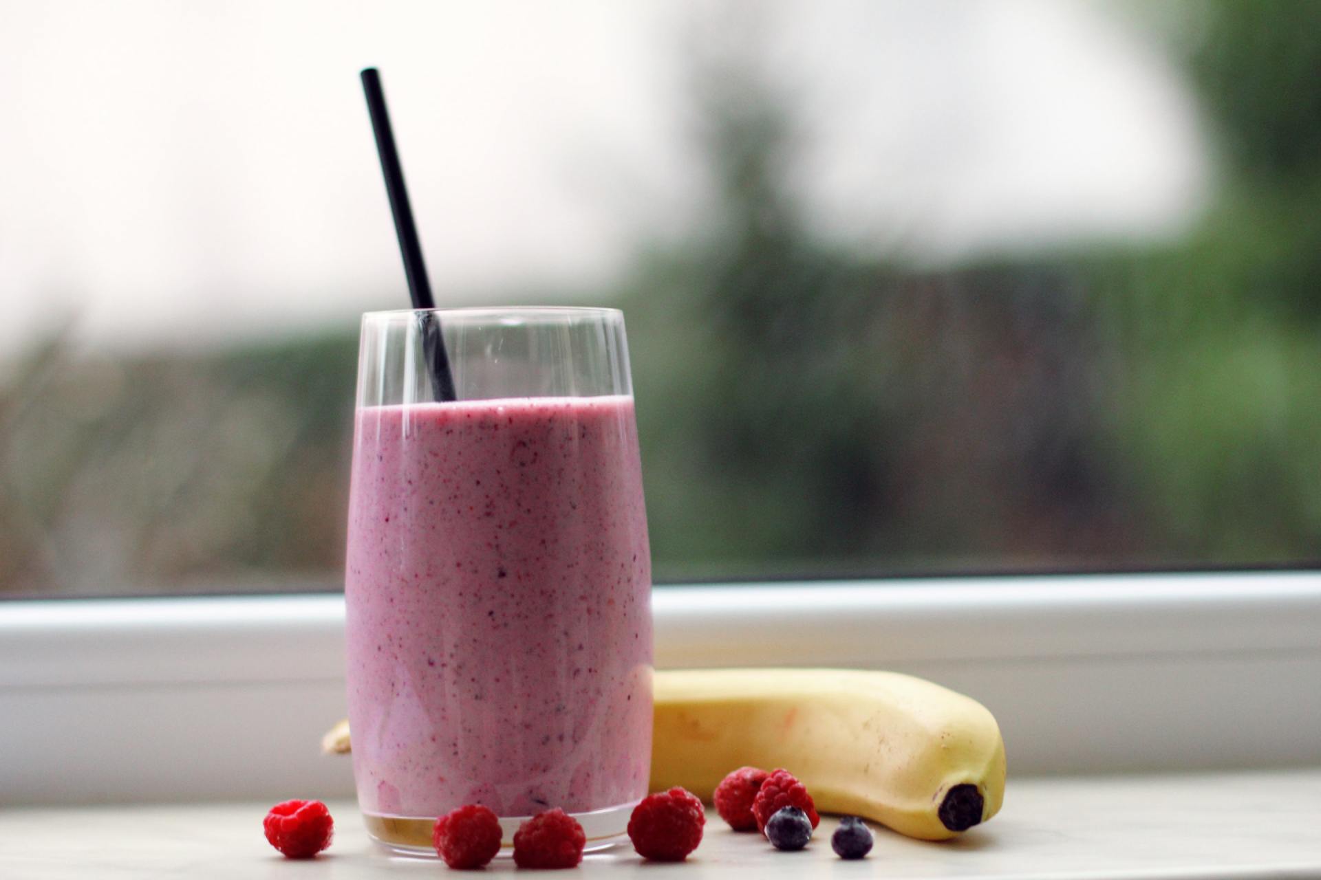 Pink smoothie in a glass with a straw, next to a banana and berries on a windowsill.