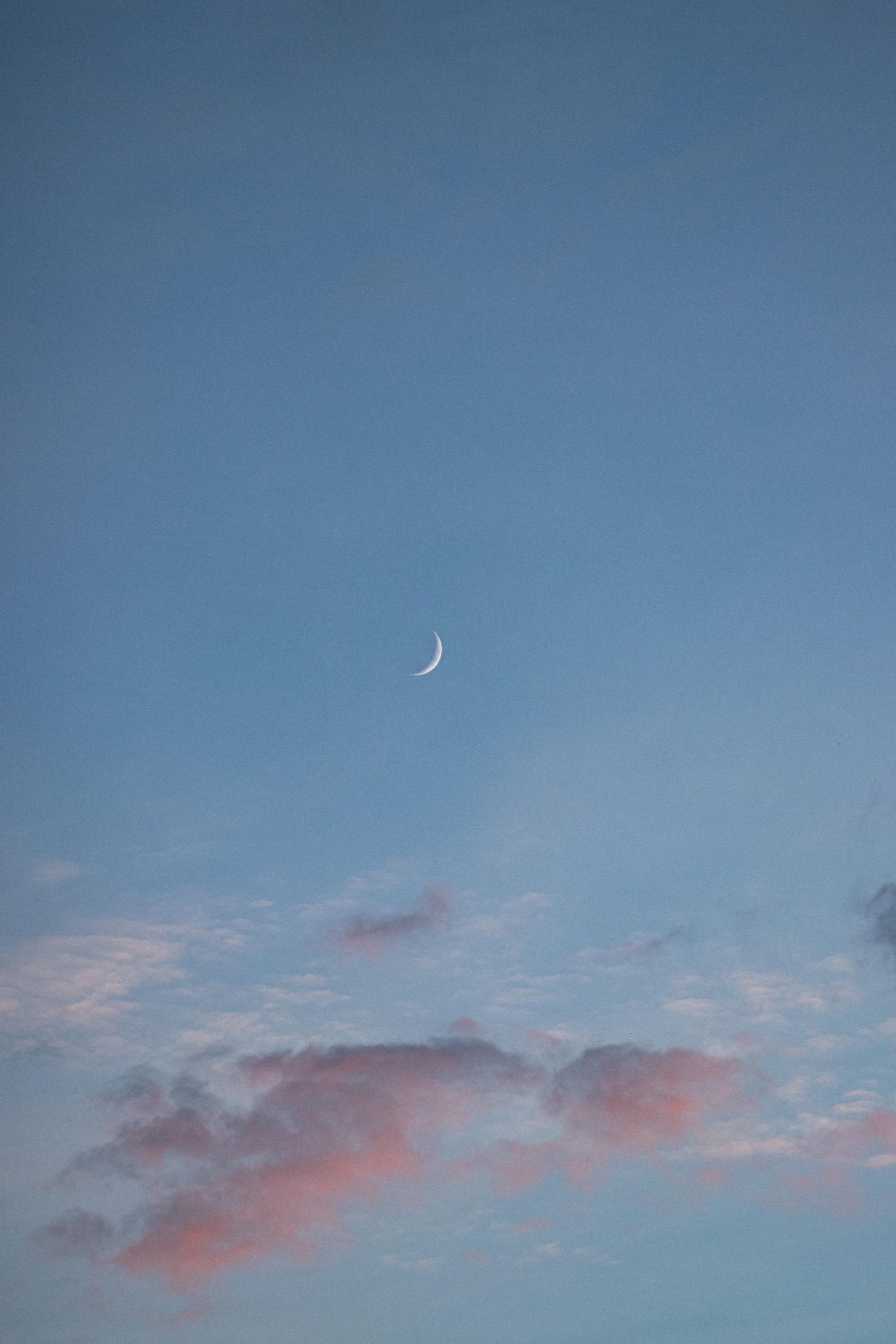 Een halve maan aan een lichtblauwe hemel met roze wolken.