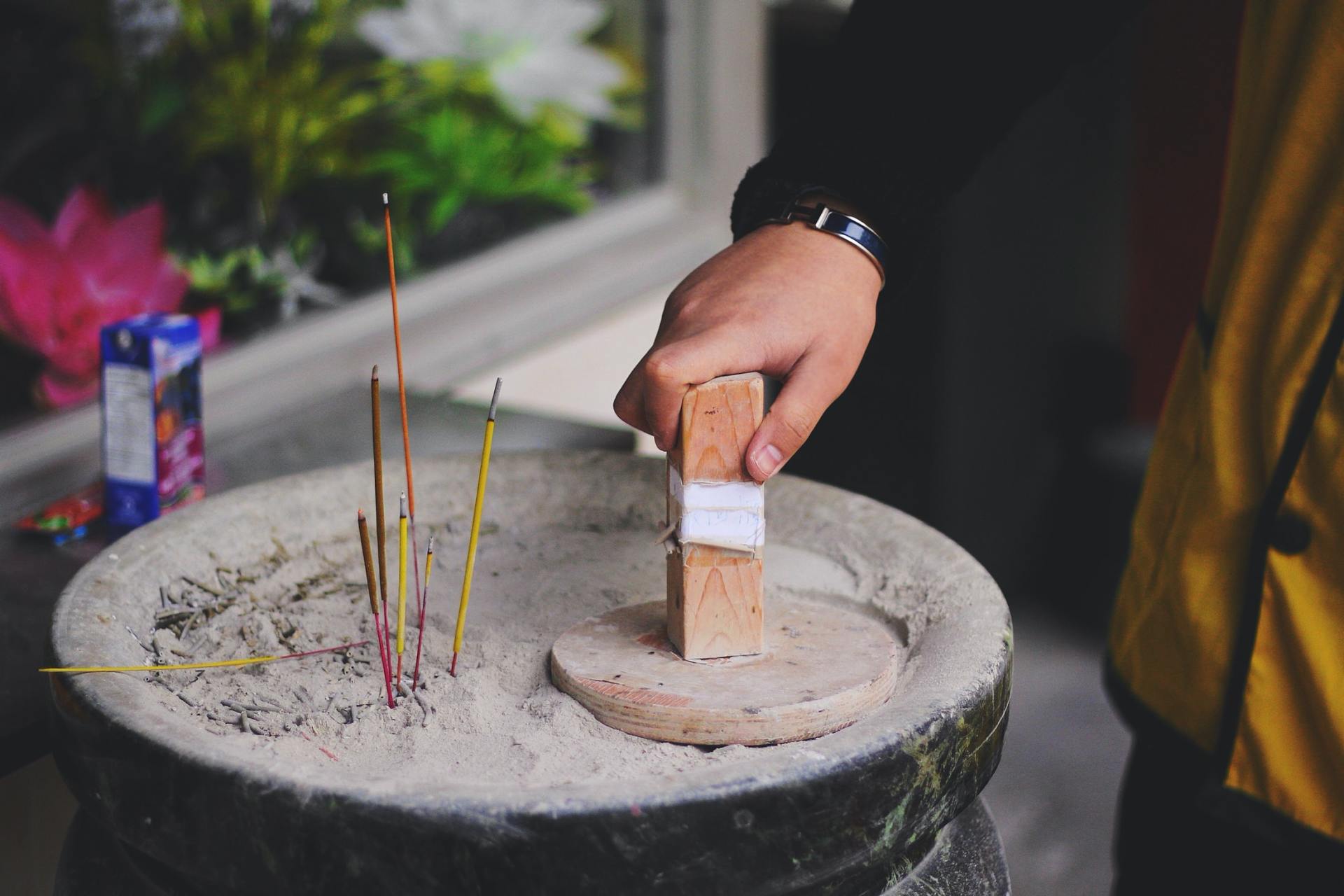 A hand places a wooden block into a stone bowl filled with sand and lit incense sticks.