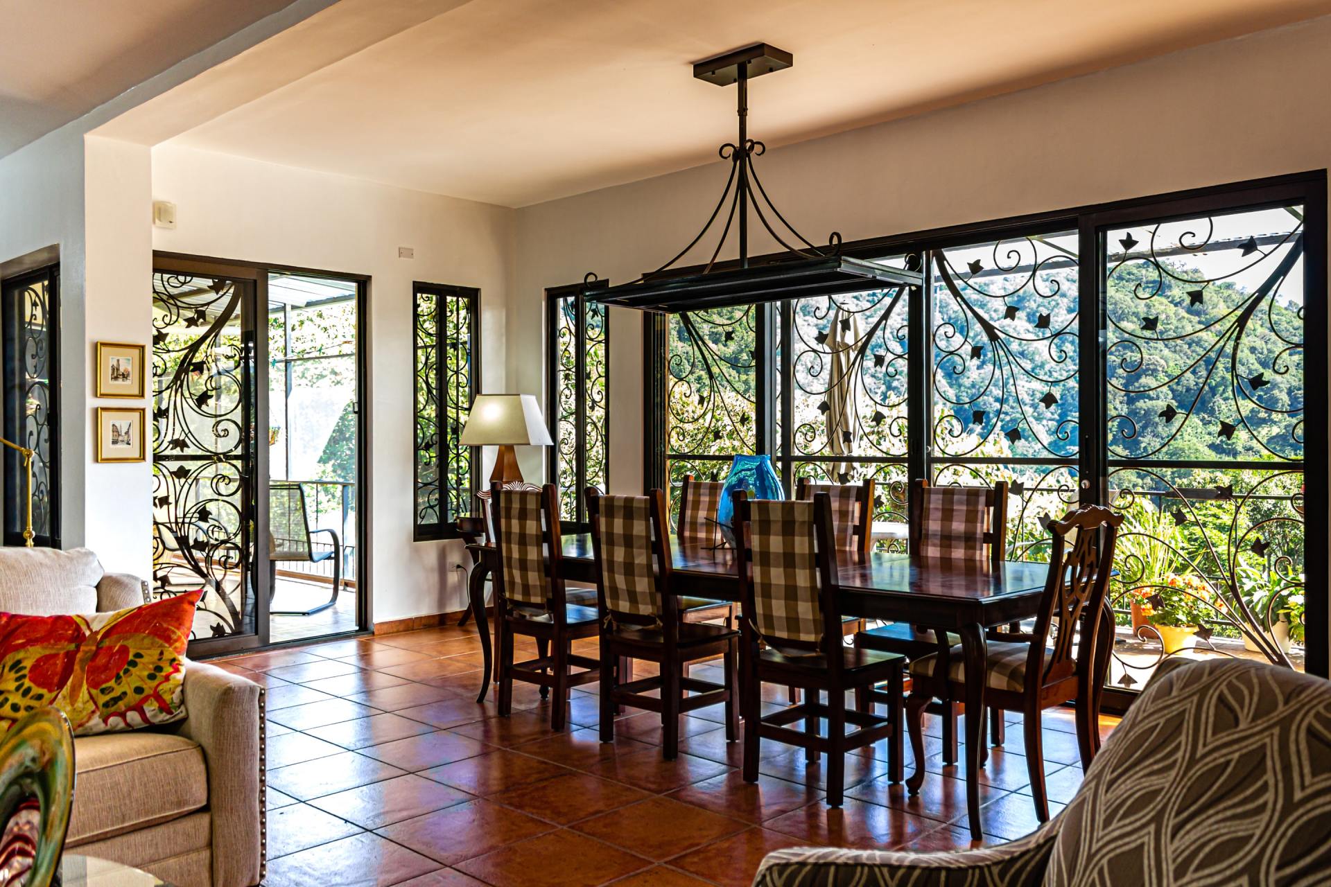 Dining room with wooden table and chairs, terracotta tile floor, and large windows overlooking greenery