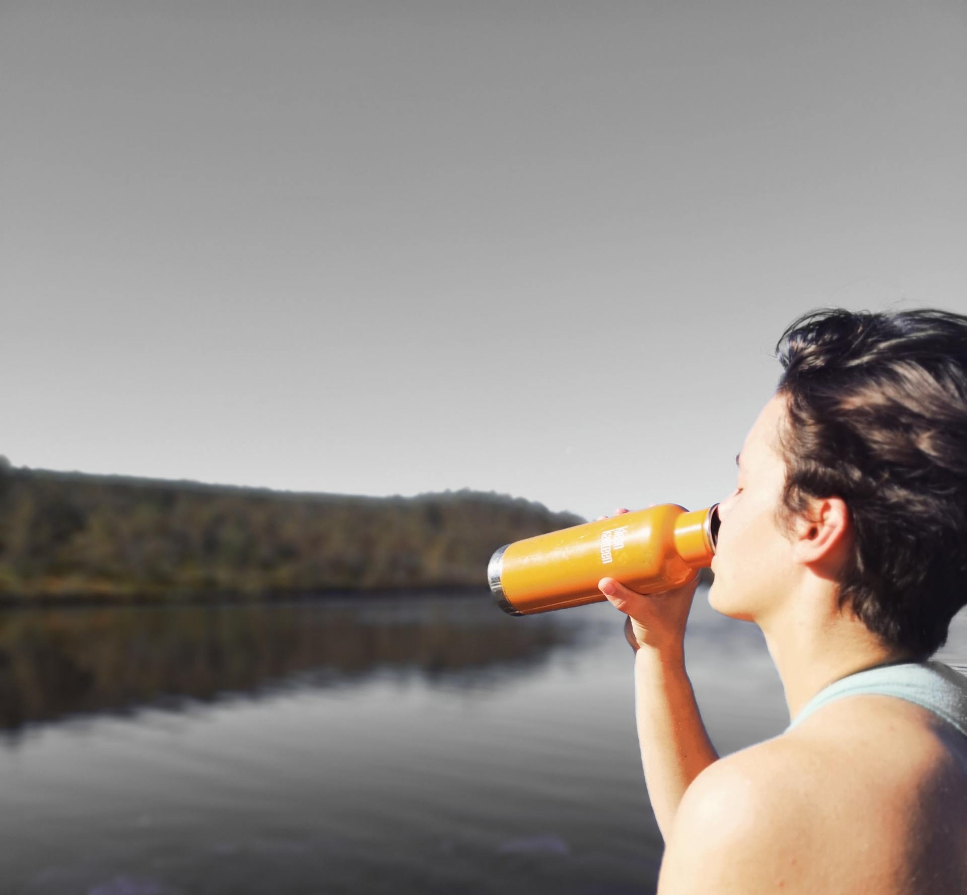 Person drinking from an orange water bottle outdoors with a lake and trees in the background.