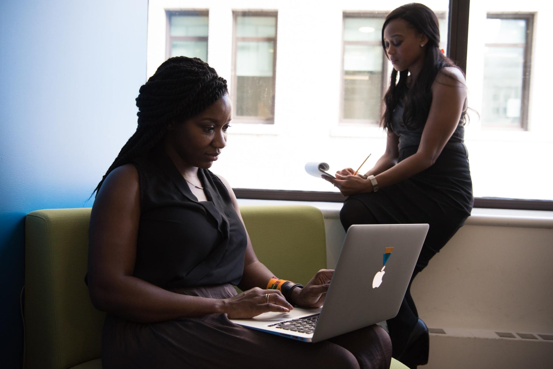 Two women are sitting on a couch using laptops.