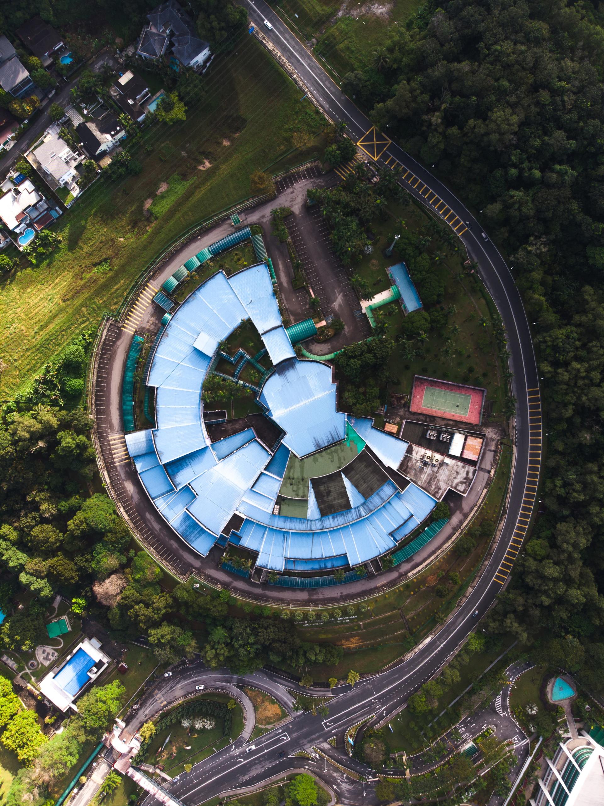 Aerial view of a circular building complex with blue roofs surrounded by roads and dense green trees.