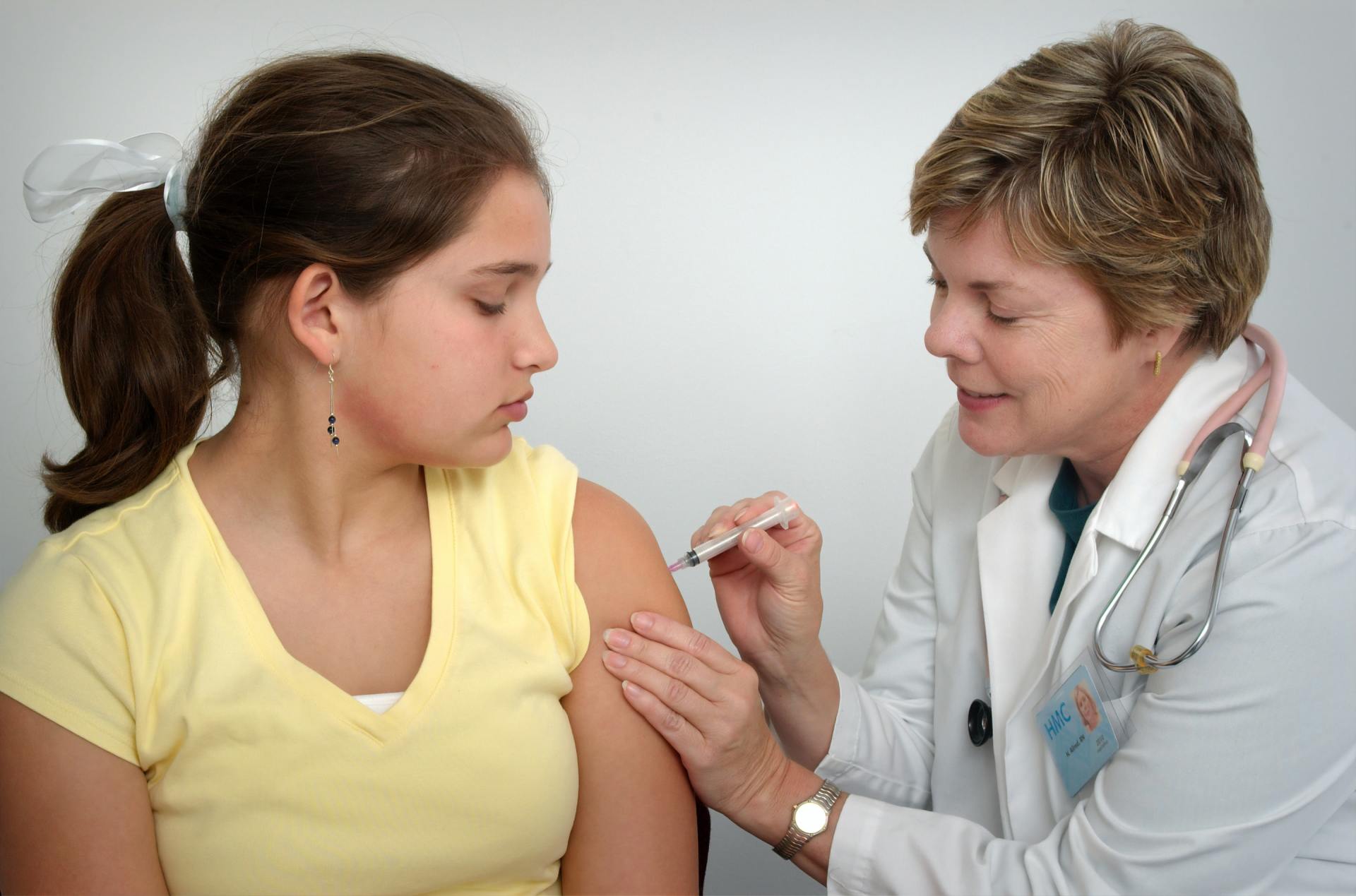 A doctor giving a patient a shot in the arm; white lab coat, yellow shirt, clinic setting.