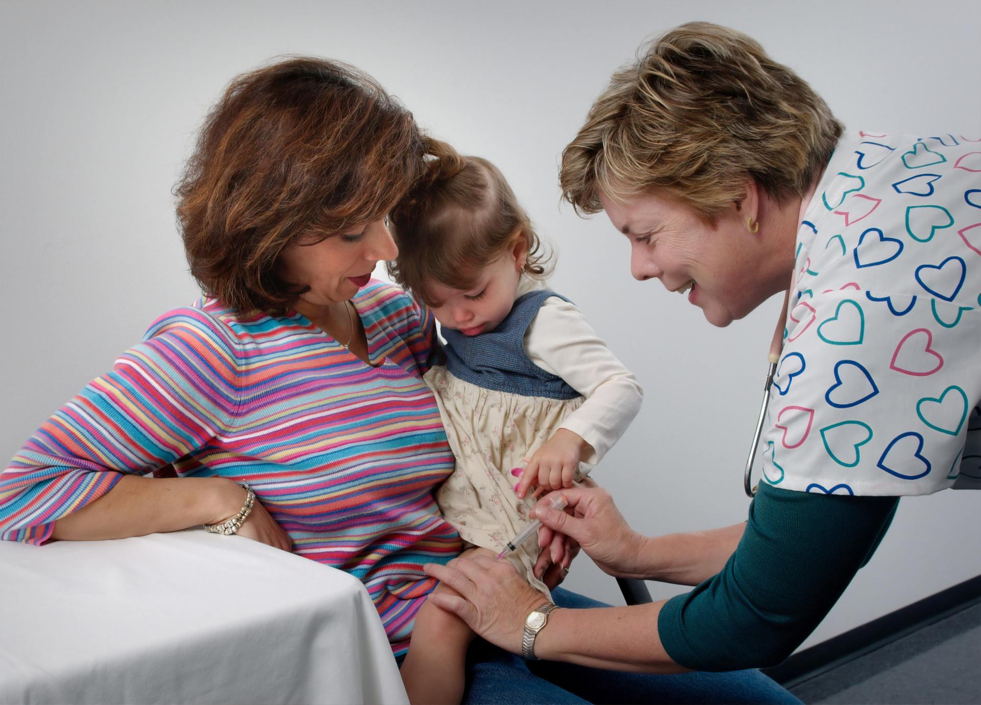 Mother comforts child during a vaccination, with a nurse; indoors, neutral background.
