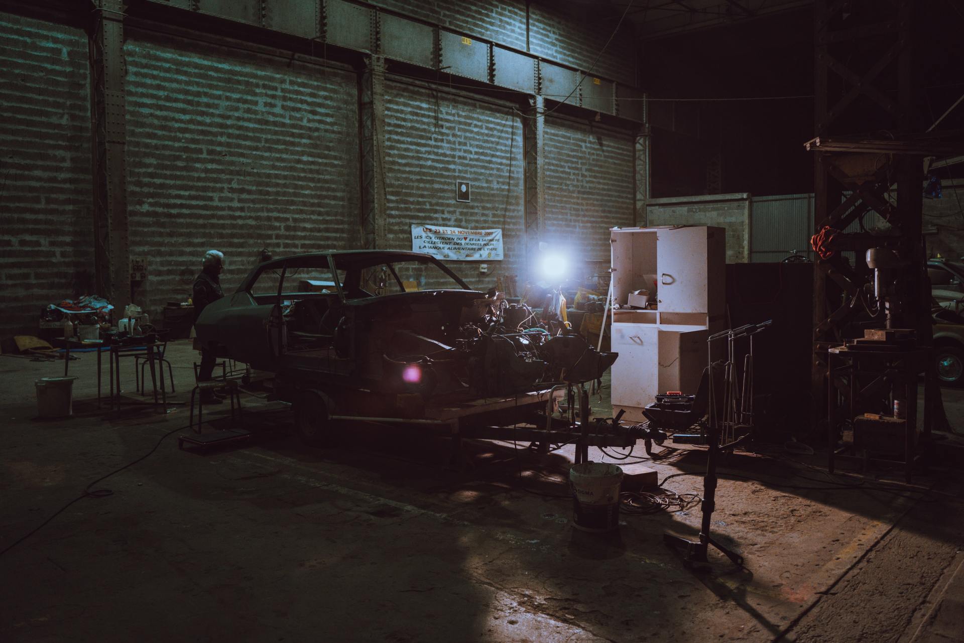 A car frame inside a dark industrial garage with a worker and bright lights.