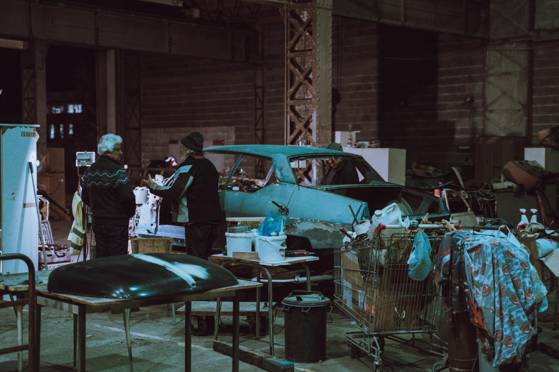 Two men work on a blue car inside a cluttered garage.