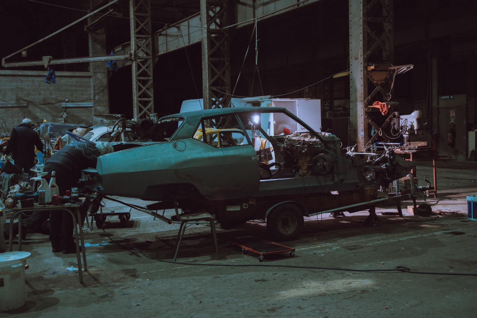 Car chassis on a trailer being worked on in a dark warehouse by two people.