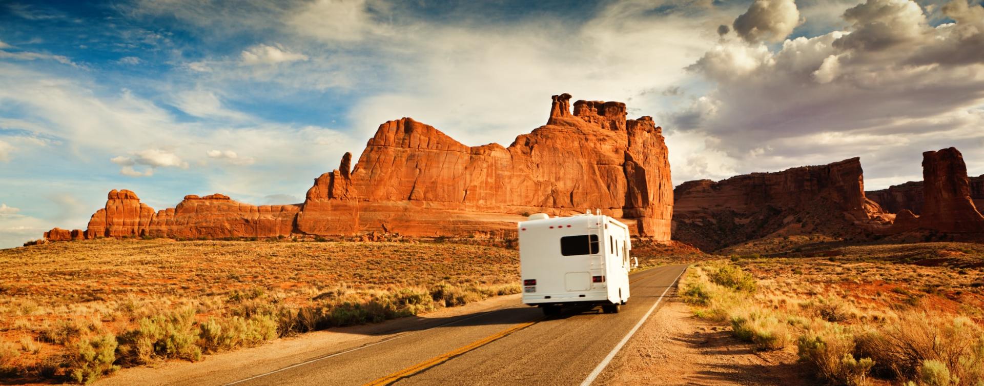 An RV drives along a winding road through red rock formations under a cloudy sky.