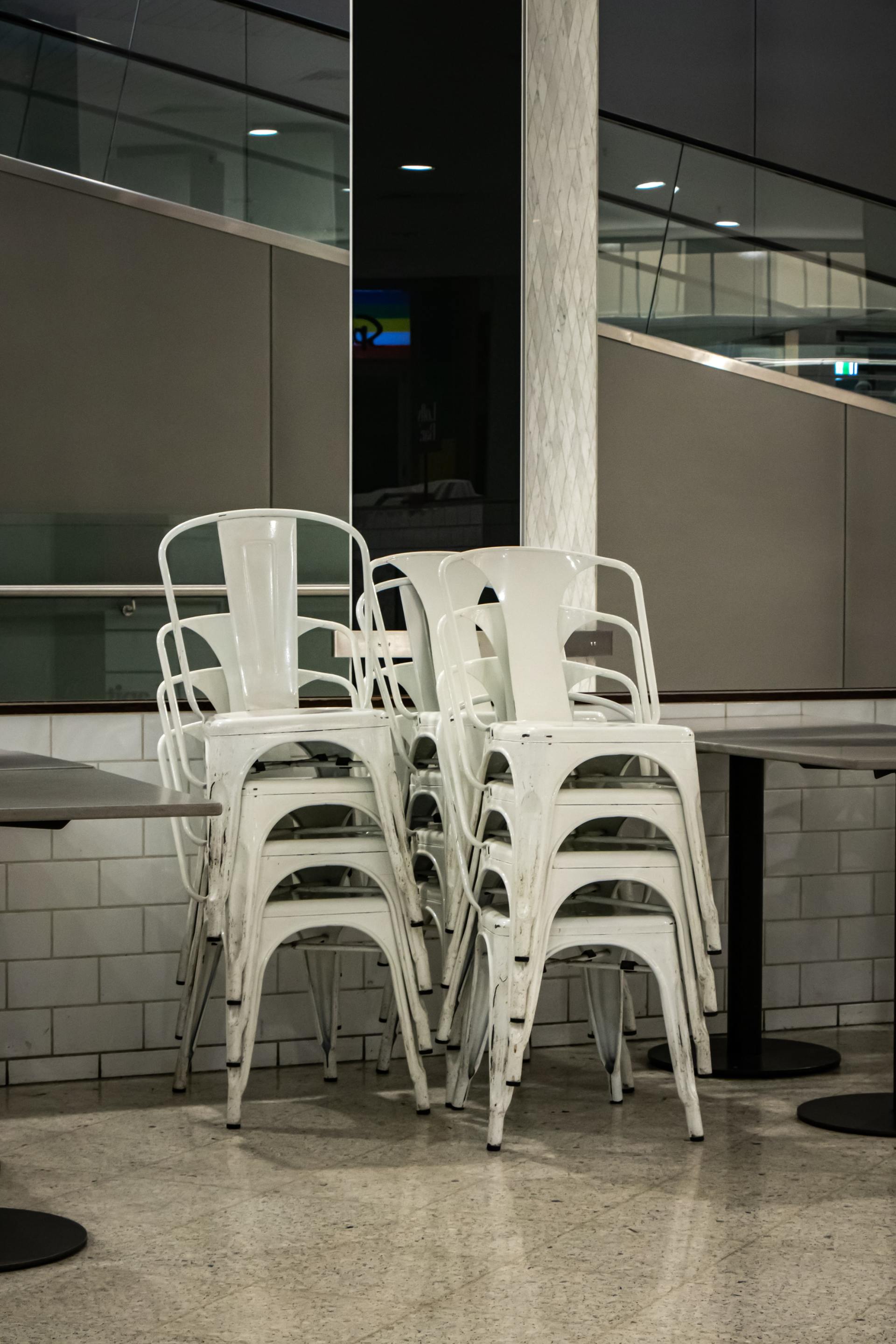 Due pile di sedie da bistrot in metallo bianco sono posizionate su un pavimento screziato, sullo sfondo di pareti piastrellate e divisori in vetro.