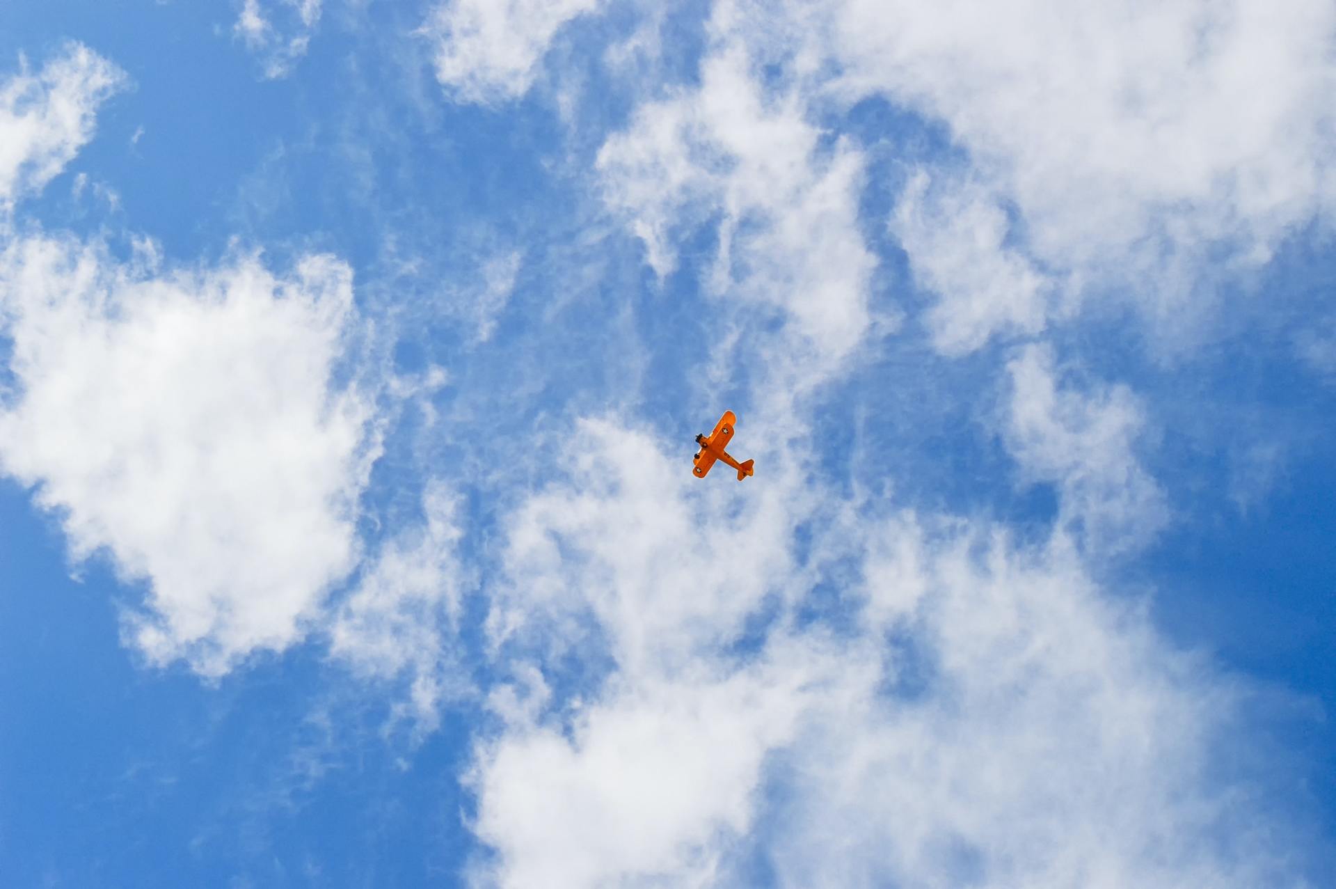 Orange biplane flying in a blue sky with white fluffy clouds.