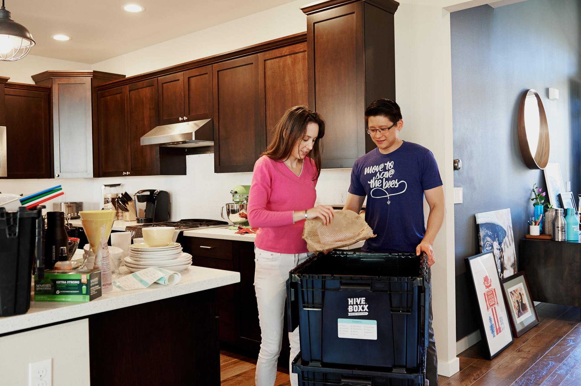A couple packing items in a kitchen, loading a container. The woman wears pink, the man a blue shirt.