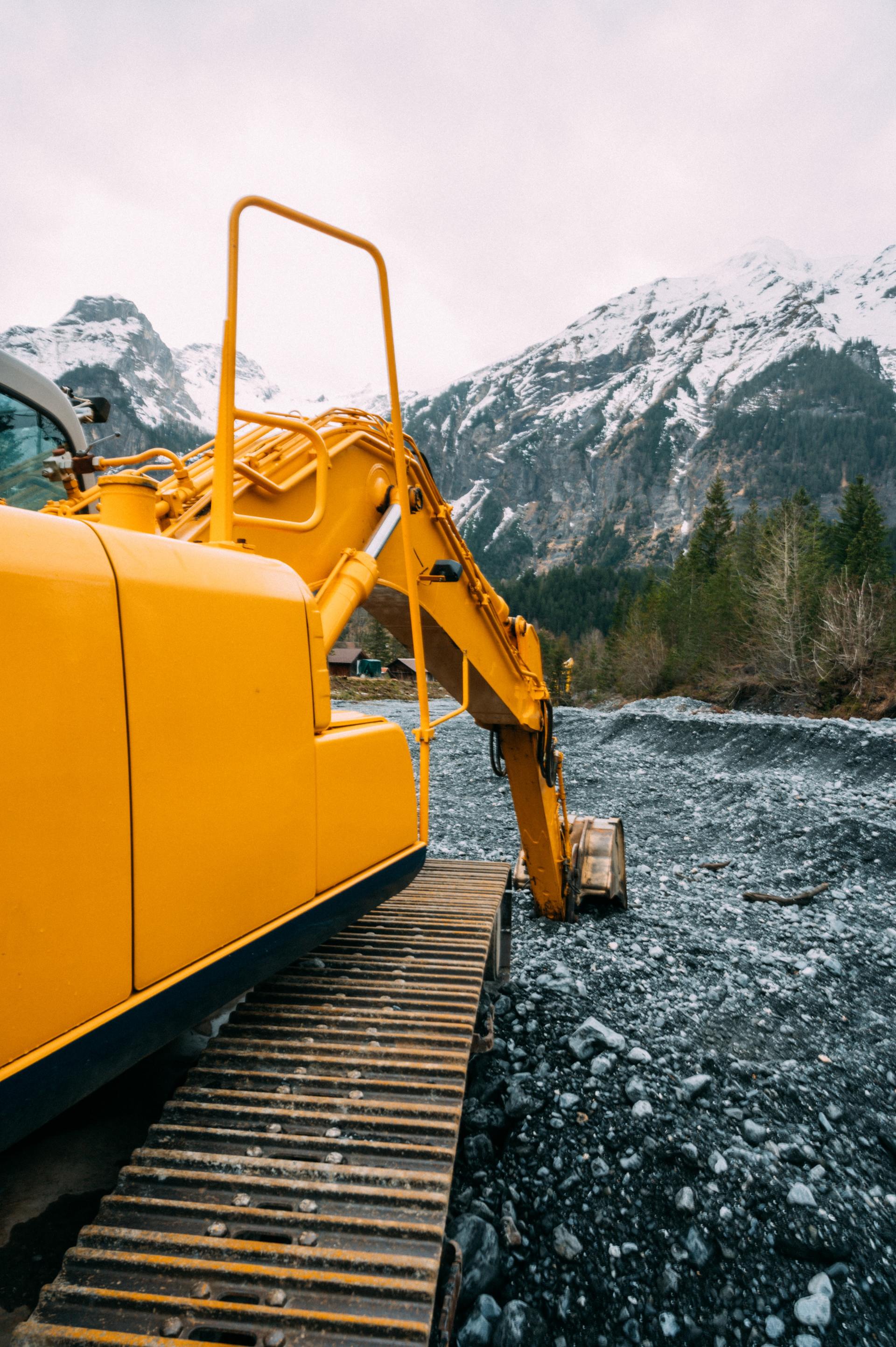 Yellow excavator on rocky terrain with snow-capped mountains in the background.
