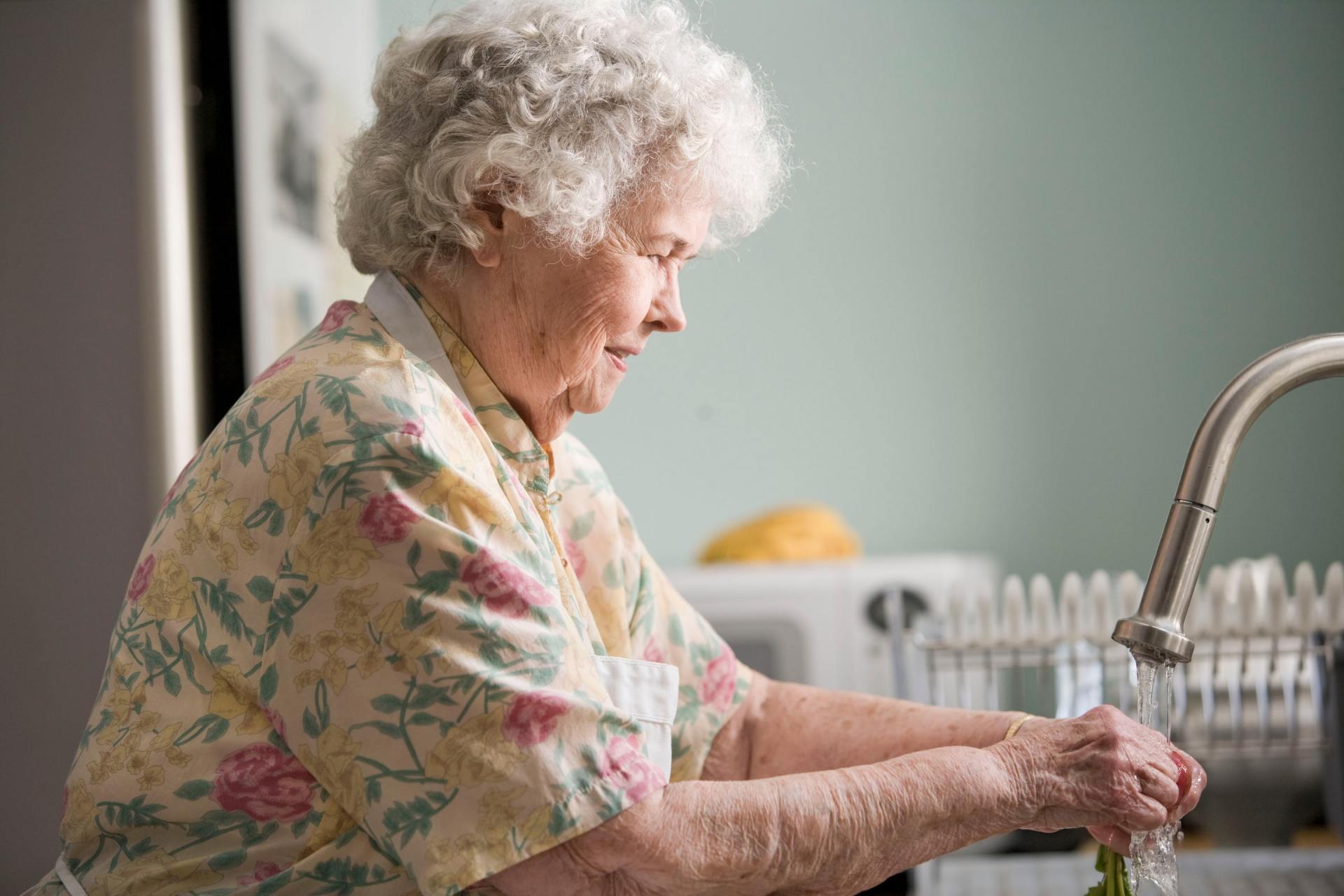 An elderly woman is washing her hands in a kitchen sink.