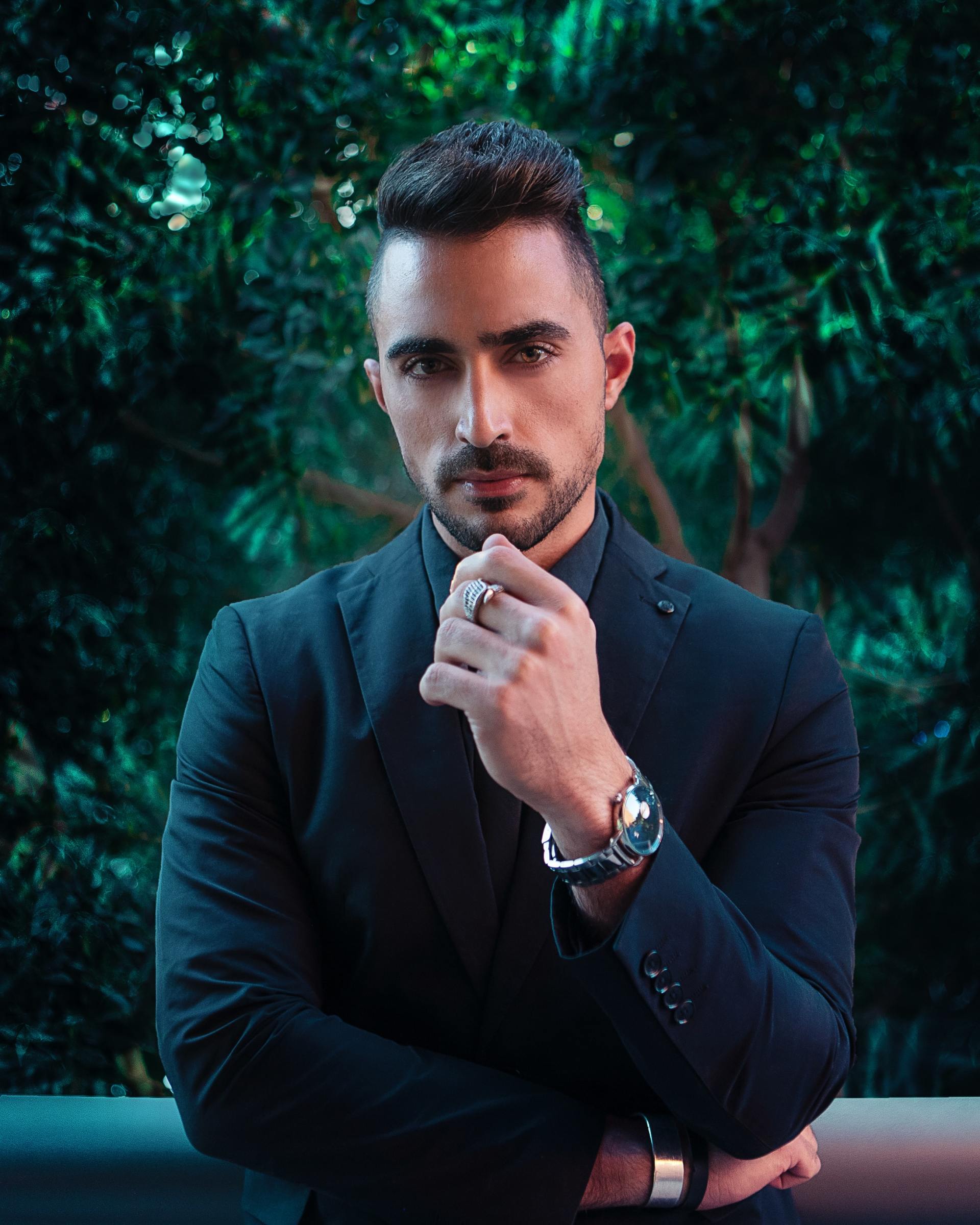 Un uomo con la barba, che indossa un abito nero e un orologio, posa con una mano al mento, sullo sfondo di un paesaggio scuro e ricco di foglie.