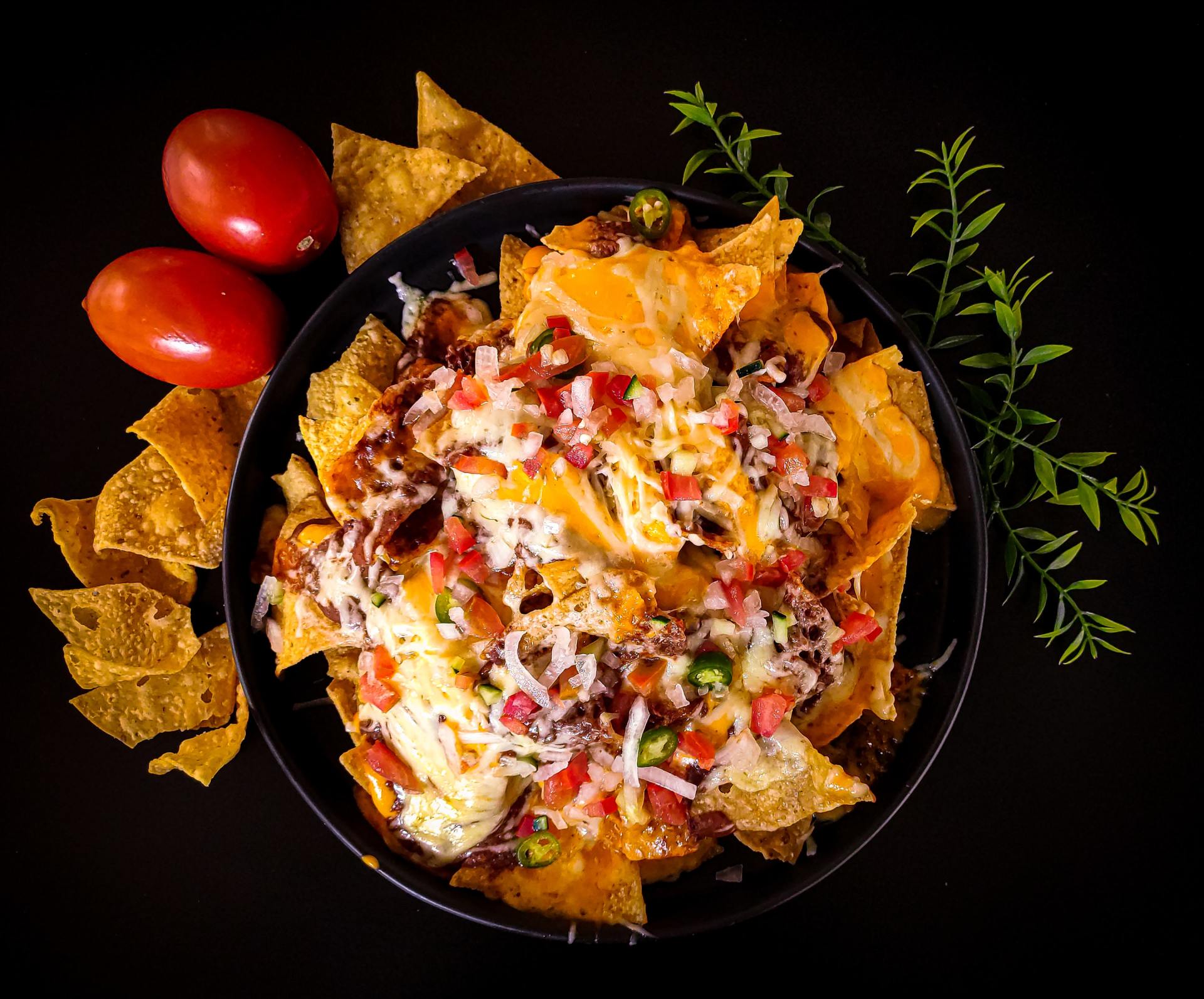 Nachos in a black bowl with cheese, toppings, tomatoes, and chips on a black surface.