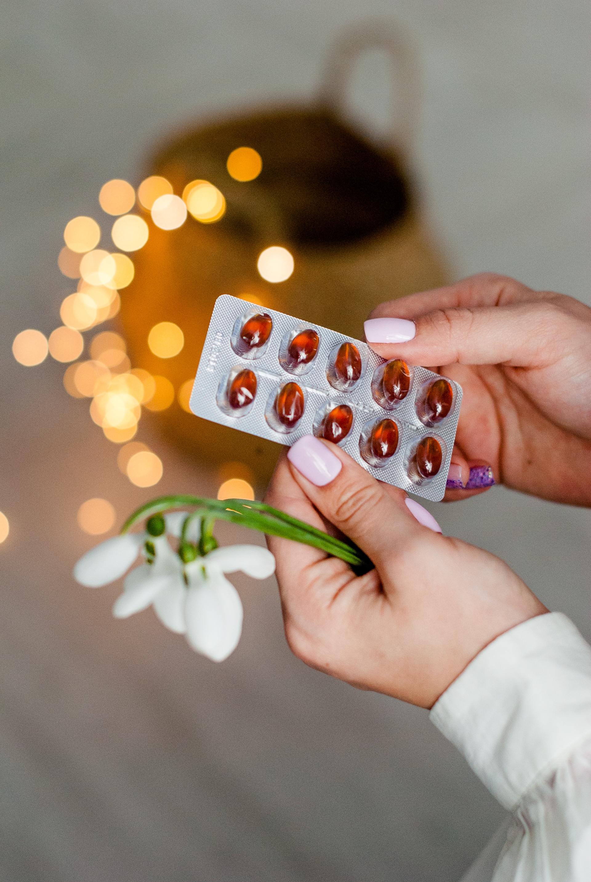 Person hält eine Blisterpackung Tabletten und ein Schneeglöckchen; verschwommener Hintergrund mit Lichtern.