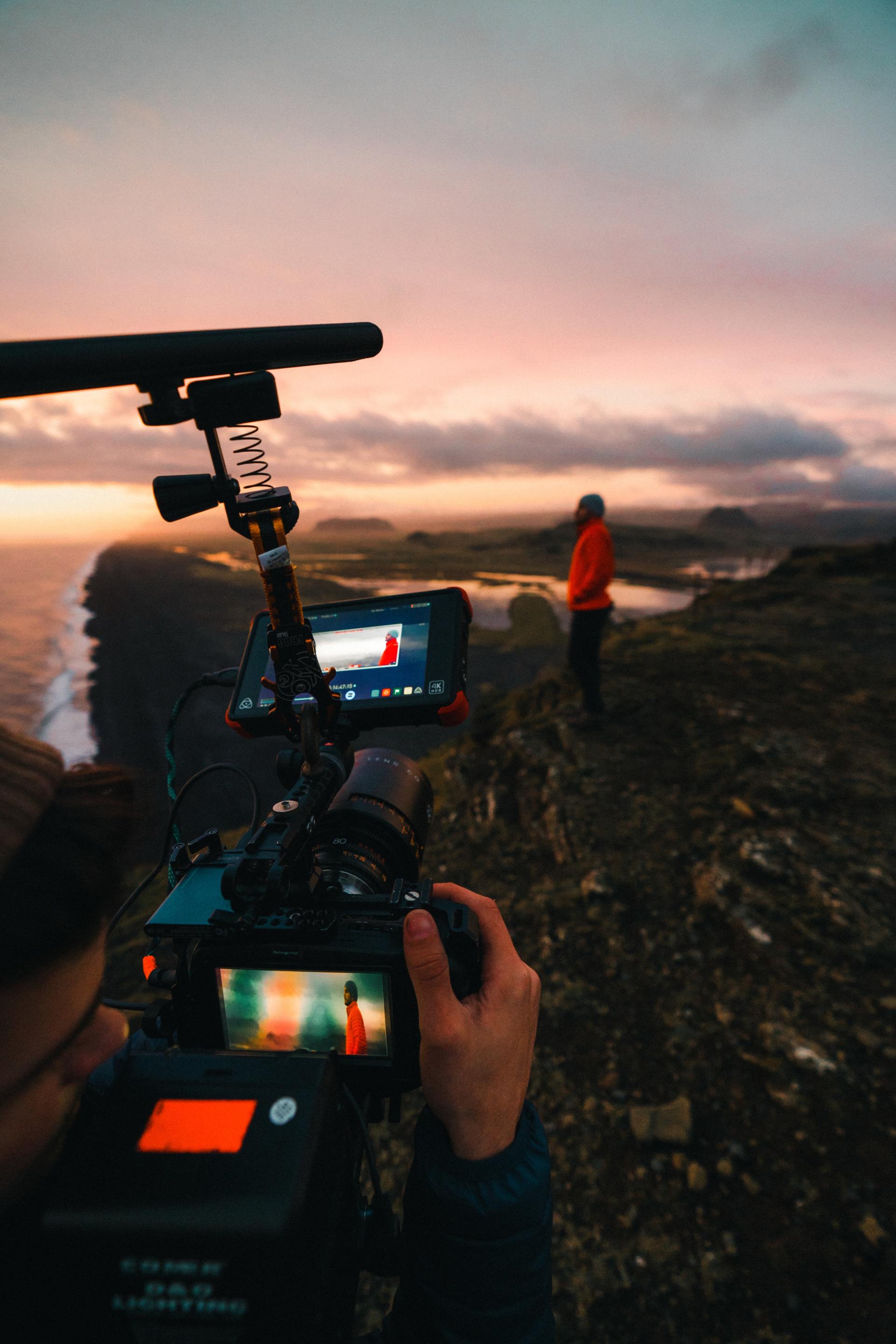 Camera filming a person in a red jacket on a rocky coastline at sunset