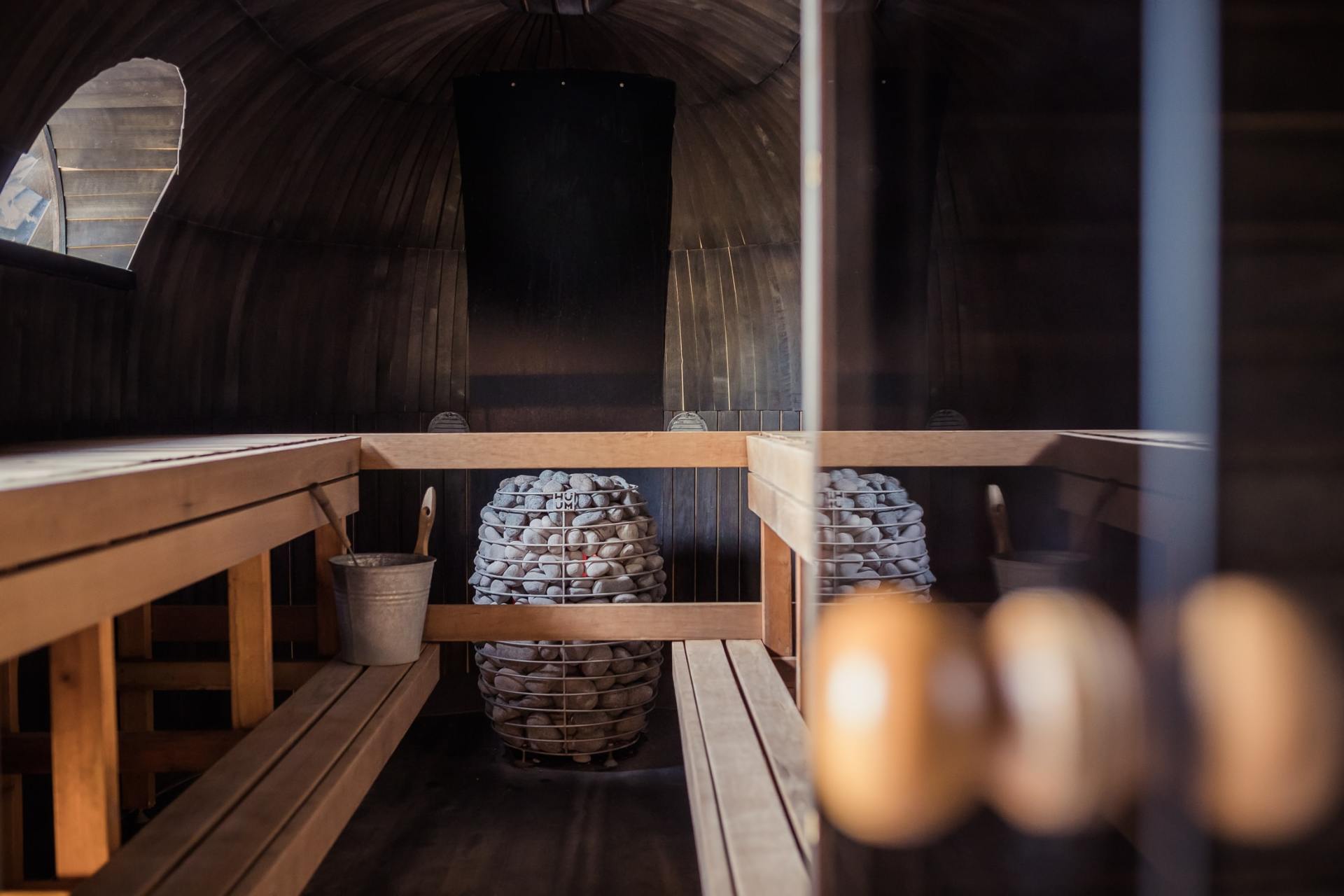 Houten sauna-interieur met banken, een emmer en een grote stenen kachel onder een gebogen plafond.