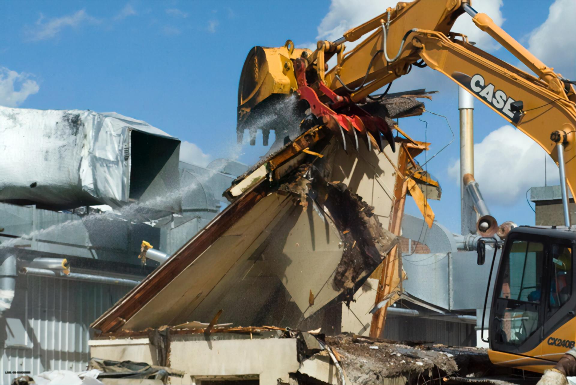 Excavator demolishing a large structure amid rubble, with dust and debris in an industrial area.