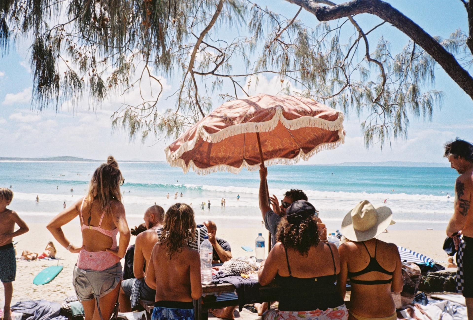 Eine Gruppe von Leuten sitzt unter einem Sonnenschirm an einem Picknicktisch am Strand.